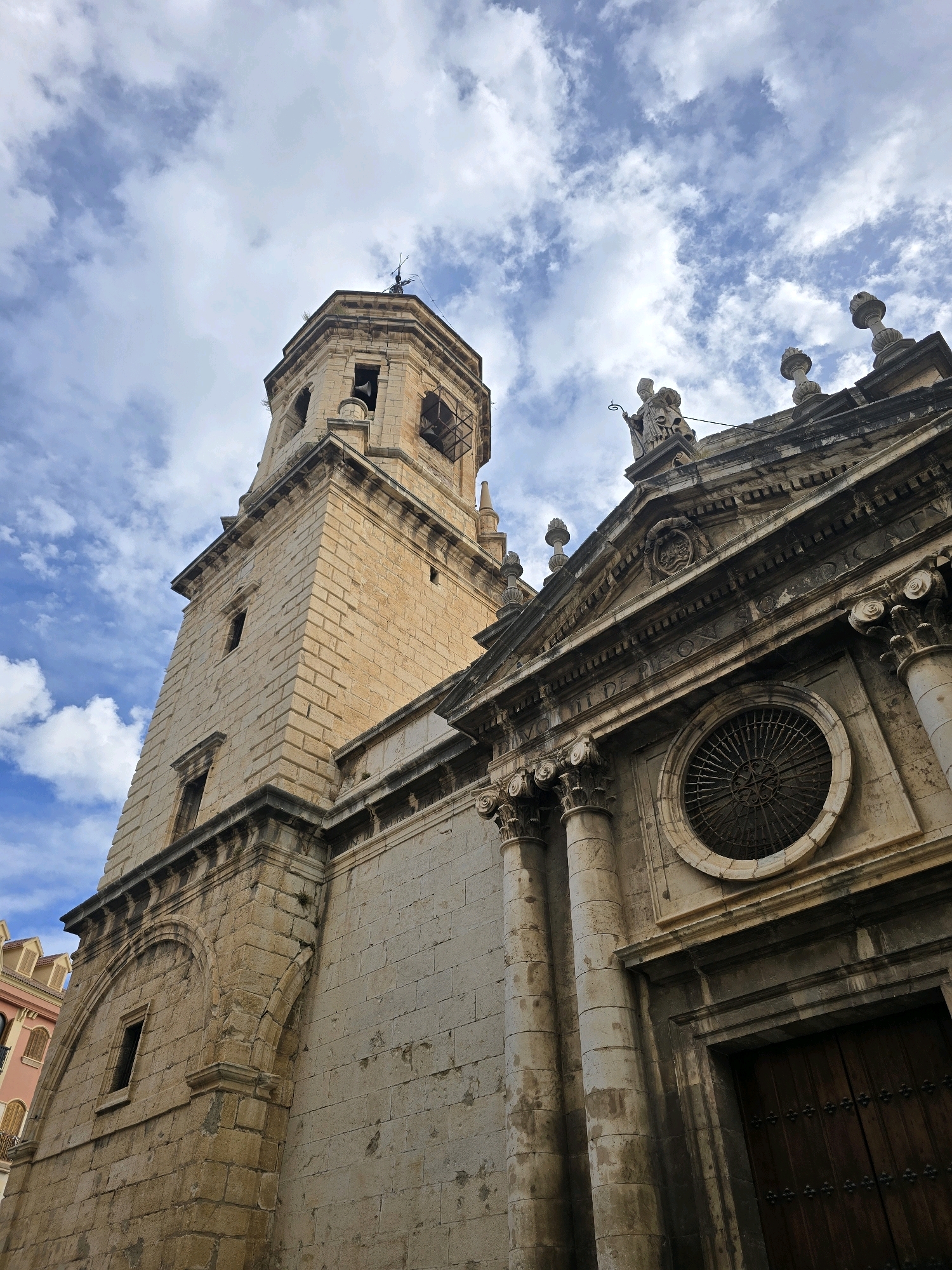 The image shows a low-angle view of the historic Cathedral of Jaén in Jaén, Spain. The cathedral's bell tower, constructed of light-colored stone blocks, dominates the left side of the frame and reaches towards a partly cloudy blue sky. The tower features several arched windows and a bell opening near its top. To the right, the ornate facade of the cathedral's main body is visible. This section is also made of stone and displays classical architectural elements, including Corinthian columns, pediments, decorative carvings, and a circular window with intricate metalwork. A statue stands atop the pediment. The rightmost part of the image shows a portion of a large, dark wooden door. The scene suggests a clear, possibly sunny day with scattered white clouds. The style of architecture indicates a significant historical religious building.