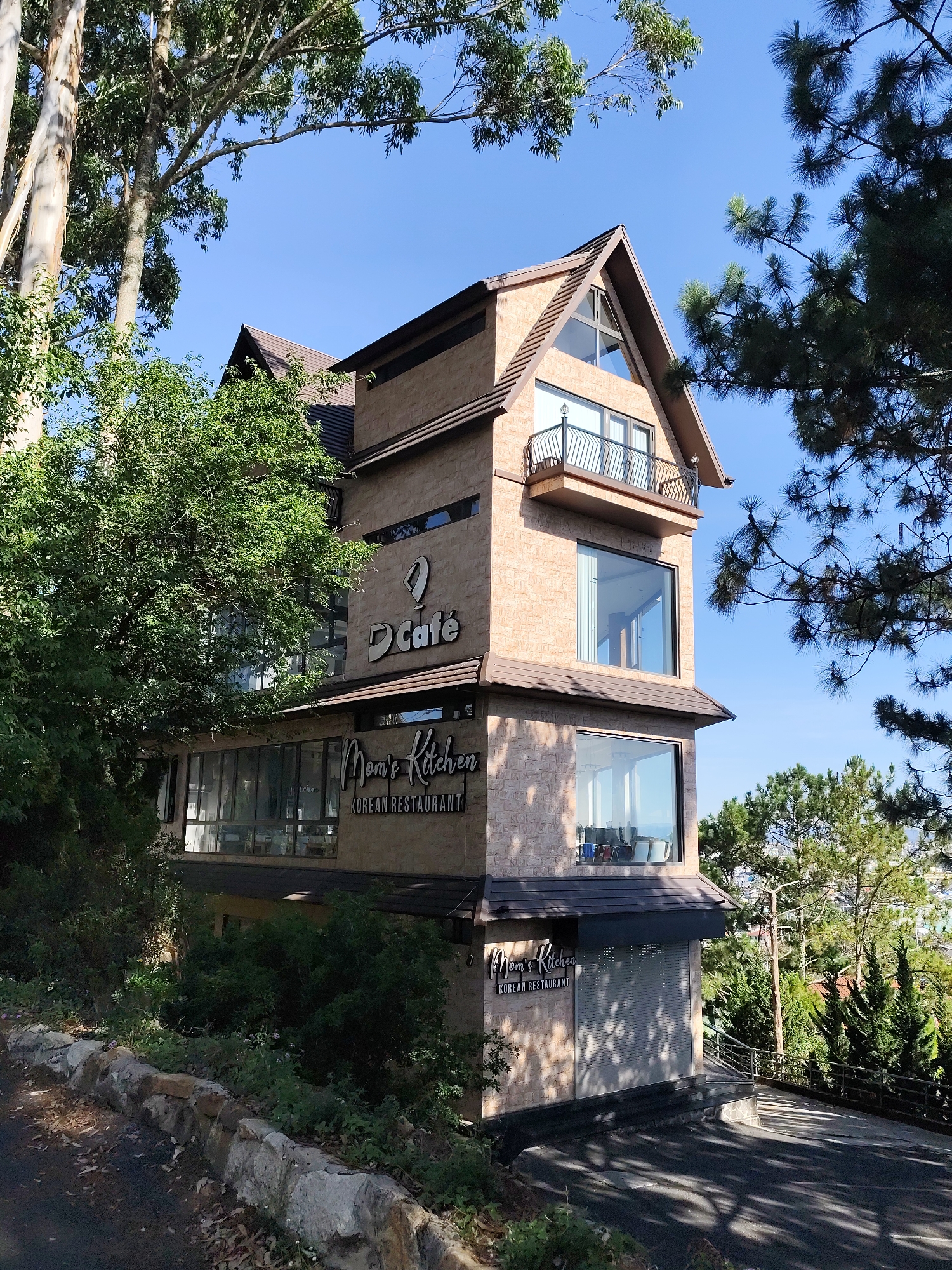 The image depicts a multi-story building housing two businesses, "D Café" and "Mom's Kitchen Korean Restaurant," in Da Lat, Vietnam. The building has a brick exterior with large windows and a small balcony on the upper floor. It is surrounded by lush greenery, including trees and bushes. The sky is clear and blue. A paved driveway leads to the building. A stone wall borders the edge of the property.