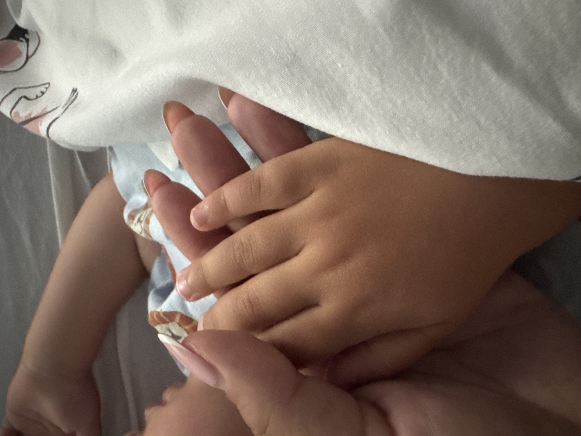 A close-up, intimate shot captures an adult's hand tenderly holding a child's hand. The larger hand, presumably an adult's, features long, manicured fingernails with a light pink base and white tips. This hand gently clasps the smaller, darker-skinned hand of a child, whose fingers are intertwined with the adult's.
The scene appears to be indoors, possibly in Sofia, Bulgaria, as indicated by the location context. Soft, indirect lighting illuminates the subjects. White fabric, likely a blanket or clothing, partially covers the top and left of the frame, showing a subtle texture and faint, light-colored line drawings of figures or objects near the upper left corner. Below the hands, glimpses of a child's clothing are visible, featuring a light blue background with a warm-toned, possibly animal or abstract, pattern in brown and white. Another arm or limb, belonging to the child, is partially visible on the left side of the frame, resting on a light-colored surface, possibly bedding. The overall impression is one of warmth, comfort, and affection between the two individuals. There is no visible text.