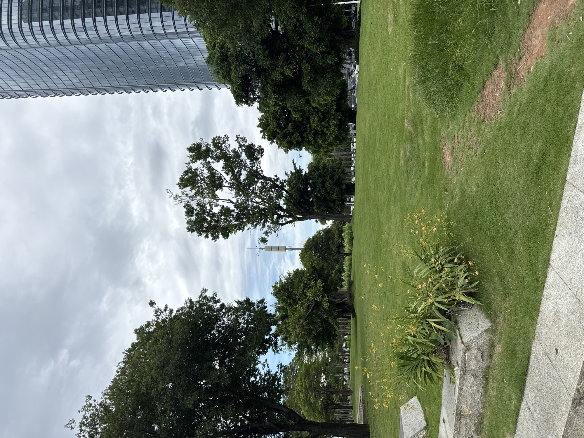 A wide shot captures a park in Tianhe District, China, under a cloudy sky. The foreground features a lush green lawn with a cluster of blooming orange flowers and a stone planter. A dirt path winds through the grass towards the right of the frame. In the mid-ground, several large trees are interspersed with a communication tower. Beyond the trees, a modern, curved skyscraper with a glass facade dominates the right side of the image. Cars are visible in the distance on a street lining the park.