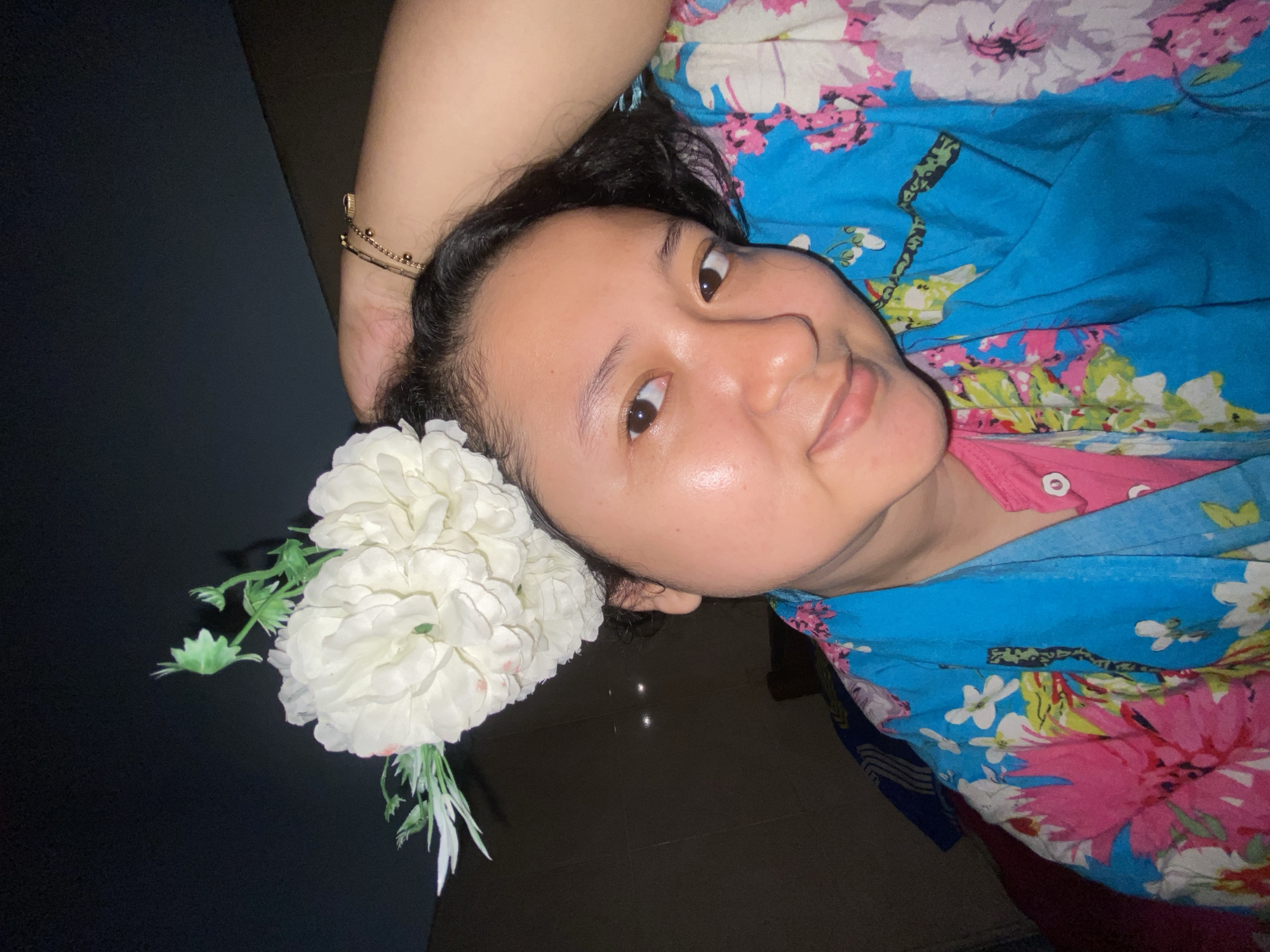 A close-up portrait, captured in Gelam, Indonesia, features an adult female looking directly into the camera with a slight smile. She has dark hair and clear skin. On the left side of her head, a prominent artificial white floral arrangement with green foliage is positioned. She wears a pink collared shirt beneath a loose, brightly patterned garment or scarf featuring large pink, white, and light blue floral designs on a blue base. Her right arm is raised, with her hand positioned behind her head. Multiple bracelets, including a thin chain with small gold spherical beads, are visible on her right wrist. The background is dark and indistinct, suggesting an indoor setting with subdued lighting, offering no specific environmental identifiers beyond general darkness.