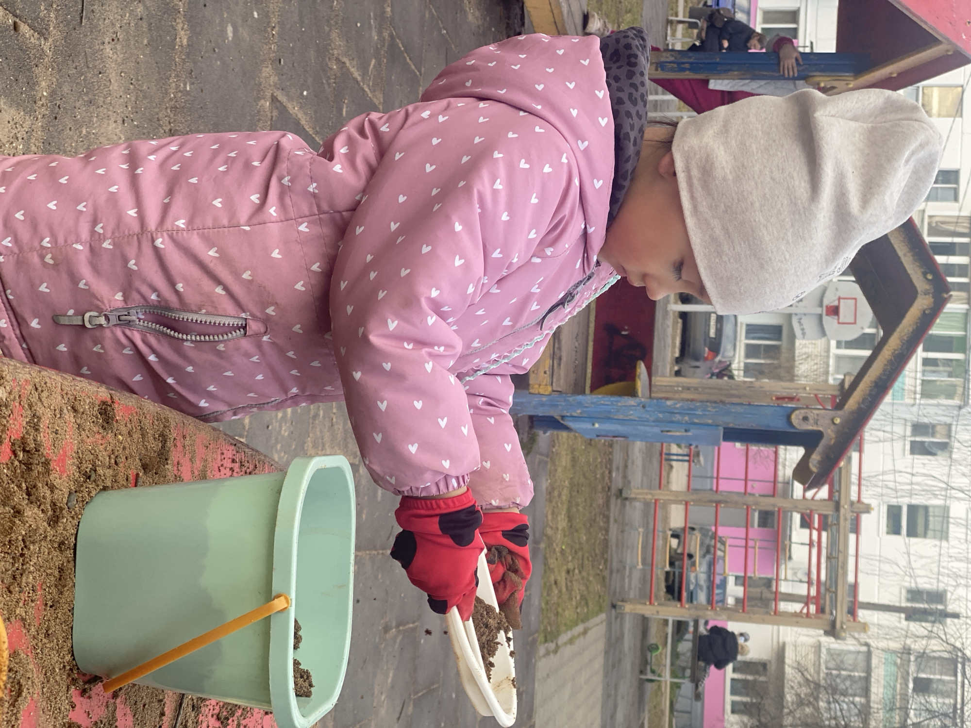 A child is playing with sand at a playground in Chaikovsky, Russia. The child, wearing a pink winter coat with white hearts, red gloves, and a light-colored hat, is holding a white plastic dish filled with sand. A light green bucket with a yellow handle is nearby, partially filled with sand. The playground equipment in the background includes climbing structures and a basketball hoop. Apartment buildings are visible beyond the playground equipment. The weather appears overcast.