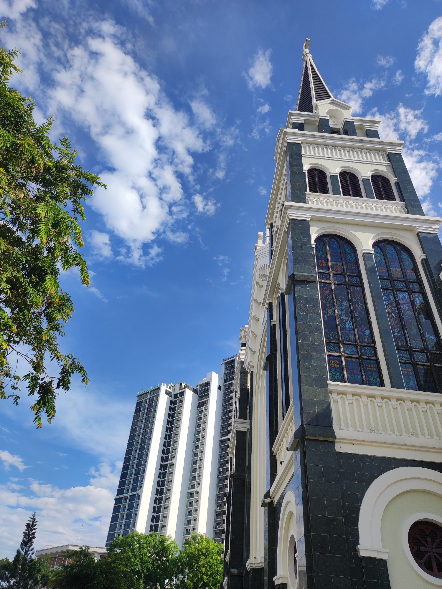 The image captures a low-angle perspective of a church tower against a bright blue sky with scattered white clouds. The church features a dark green facade with cream-colored trim and arched stained-glass windows. To the left of the church, a tall, modern white skyscraper with many windows is visible. Lush green trees are present in the foreground and at the base of the buildings. The scene appears to be a sunny day in an urban environment. This church is the Nha Trang Cathedral, a Roman Catholic cathedral in Nha Trang, Vietnam.