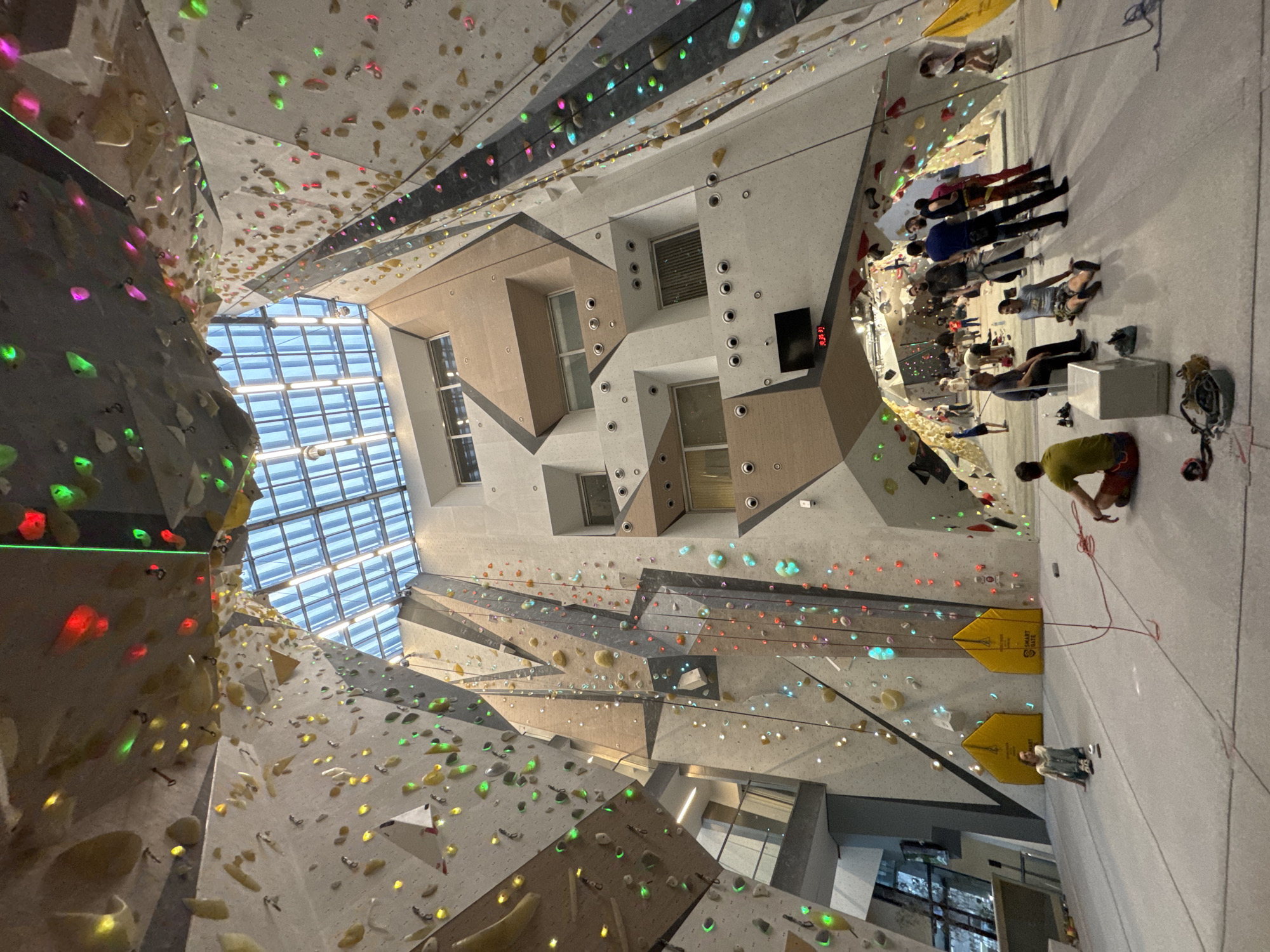The image depicts an indoor climbing gym with a high ceiling and multiple climbing walls. The location is not discernible from the image, and therefore city information cannot be provided.
The walls are covered in colorful climbing holds. Some walls have ropes attached, indicating top-roping or lead climbing routes. People are present, some sitting or standing on the floor, while others appear to be preparing to climb or resting. One person is actively being belayed.
The gym has a tall, multi-story building. A skylight allows natural light into the space. The walls have various geometric shapes and textures, giving the gym a modern look.