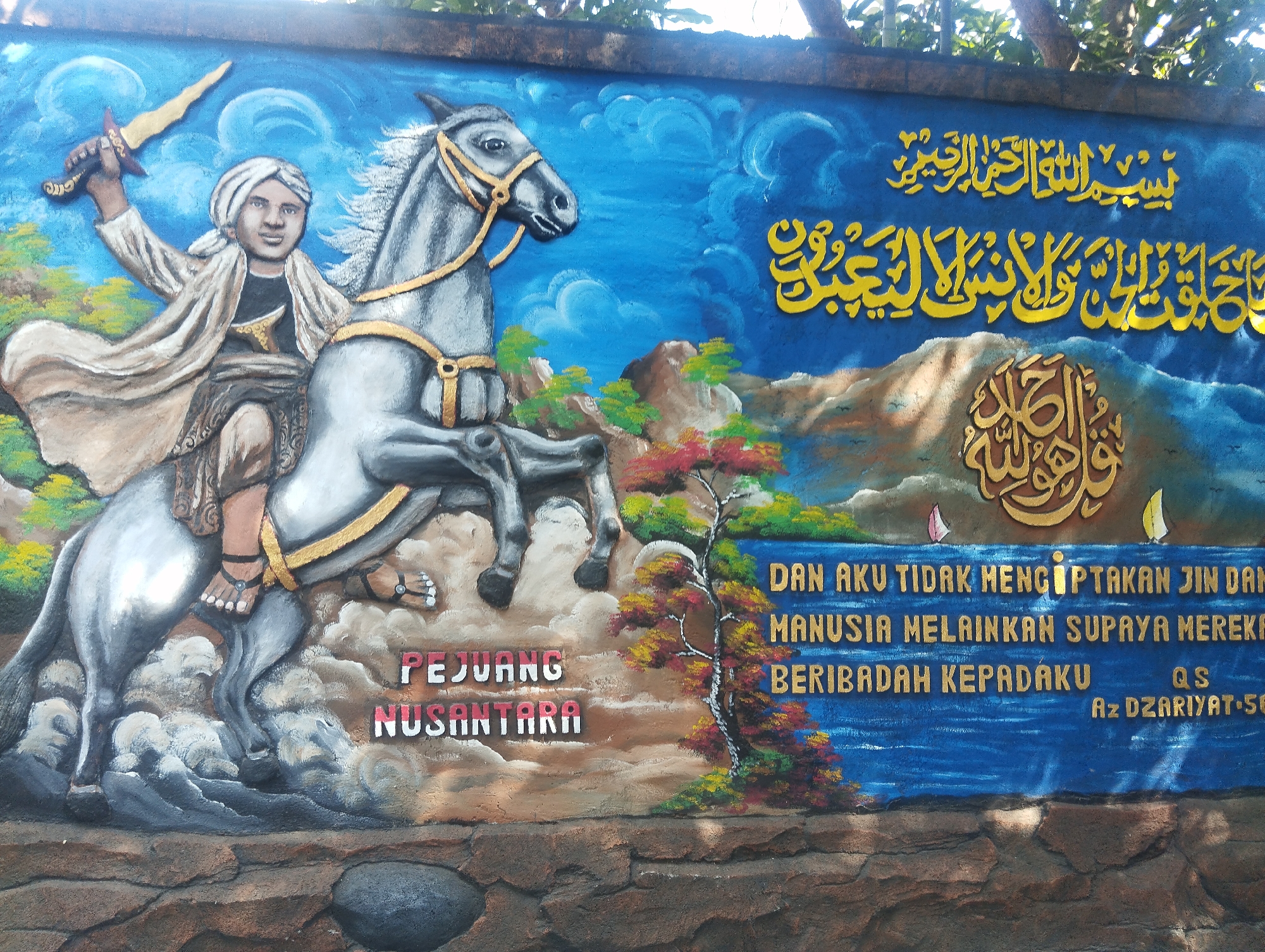 This is a photograph of a colorful mural painted on a textured wall, likely in Malang, Indonesia, given the text. The mural depicts a warrior on horseback, holding a sword aloft, against a vibrant blue sky with swirling clouds. The warrior is dressed in traditional attire, including a white turban and flowing robes. The horse is grey with a golden harness. To the right of the horse, there are mountains, a colorful tree with red and yellow leaves, and two small boats sailing on what appears to be water. In the background, there are two lines of yellow Arabic calligraphy. Below the central imagery, there are three lines of text in Indonesian: "PEJUANG NUSANTARA" (Archipelago Warrior) in red and white, followed by more yellow Arabic calligraphy, and then in blue: "DAN AKU TIDAK MENCIPTAKAN JIN DAN MANUSIA MELAINKAN SUPAYA MEREKA BERIBADAH KEPADAKU QS AZ DZARIYAT 56" (And I did not create the jinn and mankind except to worship Me, QS Az-Zariyat 56). The lower part of the wall is textured to resemble rough stone. The overall scene is dynamic and patriotic, with a religious undertone.