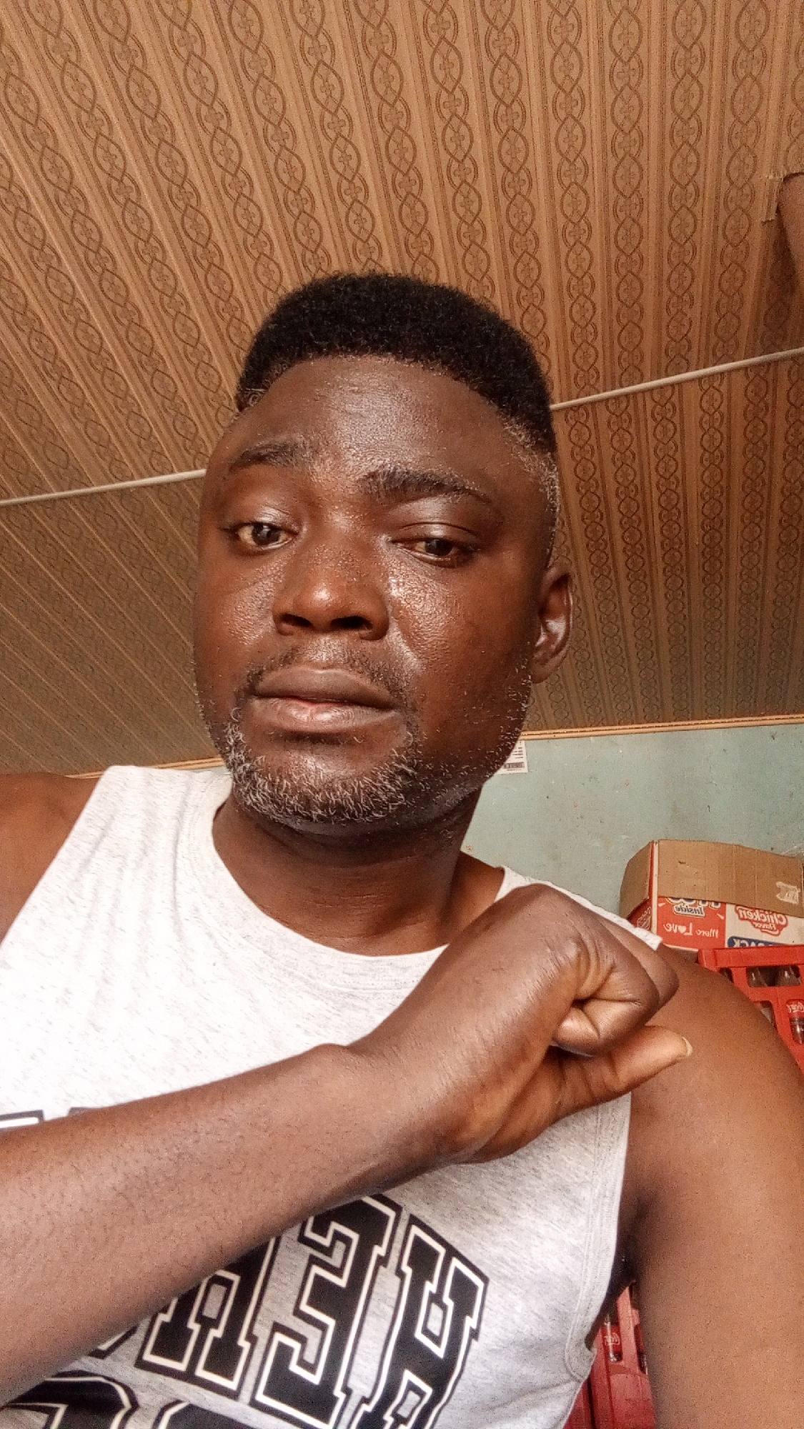 A medium shot depicts a dark-skinned man wearing a grey tank top with a bold white graphic design. The man has closely cropped black hair with grey on the sides and a salt-and-pepper beard. His skin appears to be glistening. He is resting his fist against his shoulder. The background includes a textured brown ceiling and a partial view of a pale blue wall. Boxes of "CHICKEN" crackers and red Coca-Cola crates are visible behind the man. No specific details confirm that this image was taken in the city of Narayi, Nigeria.