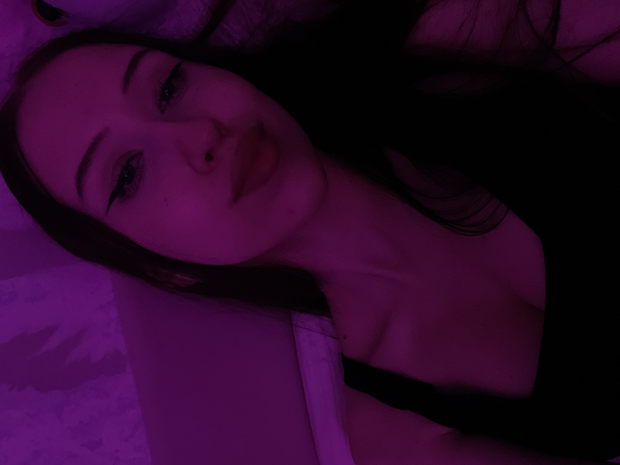 The image is a selfie of a young woman with long dark hair and winged eyeliner, taken in what appears to be a dimly lit indoor setting with purple ambient lighting. The background is textured and shows some shadow play. The woman is wearing a black tank top, and her skin tone is illuminated by the purple light. The overall impression is that of a casual, personal photograph. There is no discernible information within the image that would indicate its location in Votkinsk, Russia.