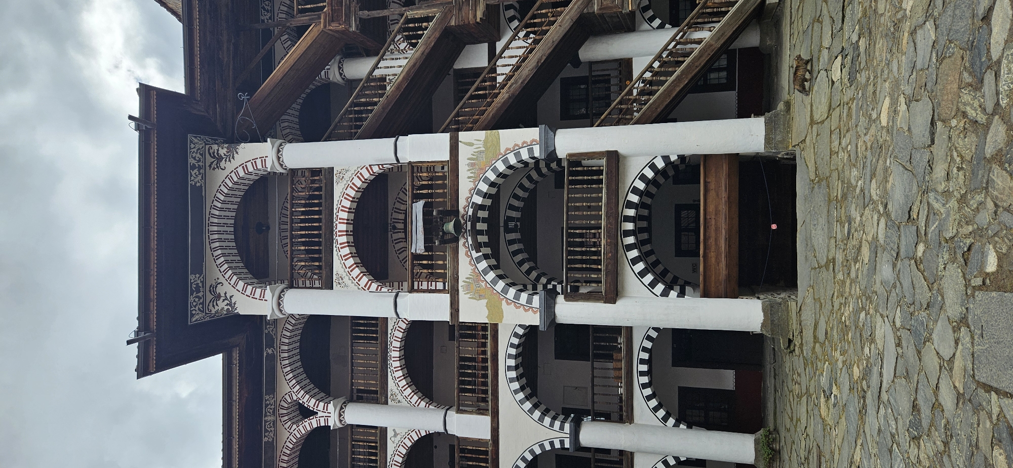 The image displays a multi-story section of the Rila Monastery in Rilski manastir, Bulgaria, specifically a portion of its residential quarters facing an inner courtyard, under an overcast sky.
The scene is dominated by the monastery's distinctive architecture. The building facade features a series of three visible levels above ground, constructed with prominent white columns supporting numerous arched openings. The arches on the ground floor are decorated with alternating black and white horizontal stripes, while those on the upper floors exhibit alternating red and white horizontal stripes. Each arched opening typically frames a wooden balcony with turned balusters. Mural paintings, depicting architectural landscapes and ornate floral motifs, are visible on the white wall sections between the arched windows, particularly above the lower-level arches on the right.
A large, antique-style brass bell is suspended from a wooden beam within one of the first-floor arches on the right side. Below this, a dark, recessed entrance or passage is visible, above which a "WC" sign is discreetly placed. A thin string with a small, red, rose-shaped object is strung across the entrance of this passage.
On the far right, a rustic, multi-flight wooden staircase with wooden balustrades ascends diagonally, providing access to the upper levels of the monastery. The building is topped with dark wooden eaves and a dark roofline, against which a grey, cloudy sky is visible.
The ground surface consists of irregular grey stone paving. A single domestic tabby cat, with brown and black striped fur, walks from right to left across this paving in the lower right quadrant of the image. No other individuals or animals are present.