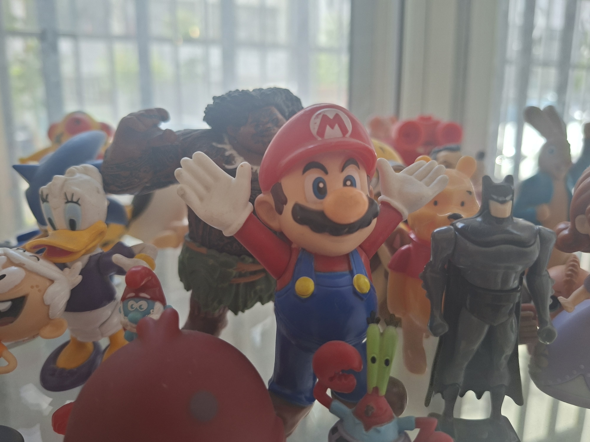 A collection of various toy figurines, including Mario, Maui, Batman, Mr. Krabs, Donald Duck, a Smurf, Winne the Pooh, and other indiscernible characters, are clustered together indoors. The setting appears to be near a window, possibly in a home in Tel-Aviv, Israel, based on the blurred urban backdrop suggesting a residential or commercial area with trees visible through the glass. There are no people visible in the image.