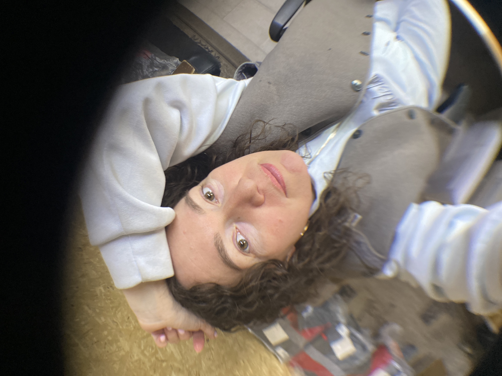 A woman with curly brown hair and fair skin looks up at the camera, her face slightly angled. She is wearing a white hooded sweatshirt and a gray vest over a light blue collared shirt. Her hands are near her face. The background is blurred, showing glimpses of a floor, a chair, and some other objects. The overall composition appears to be a selfie taken from a low angle, looking up. There is no evidence to confirm the location is in the city of Ol'khovskoye, Russia.