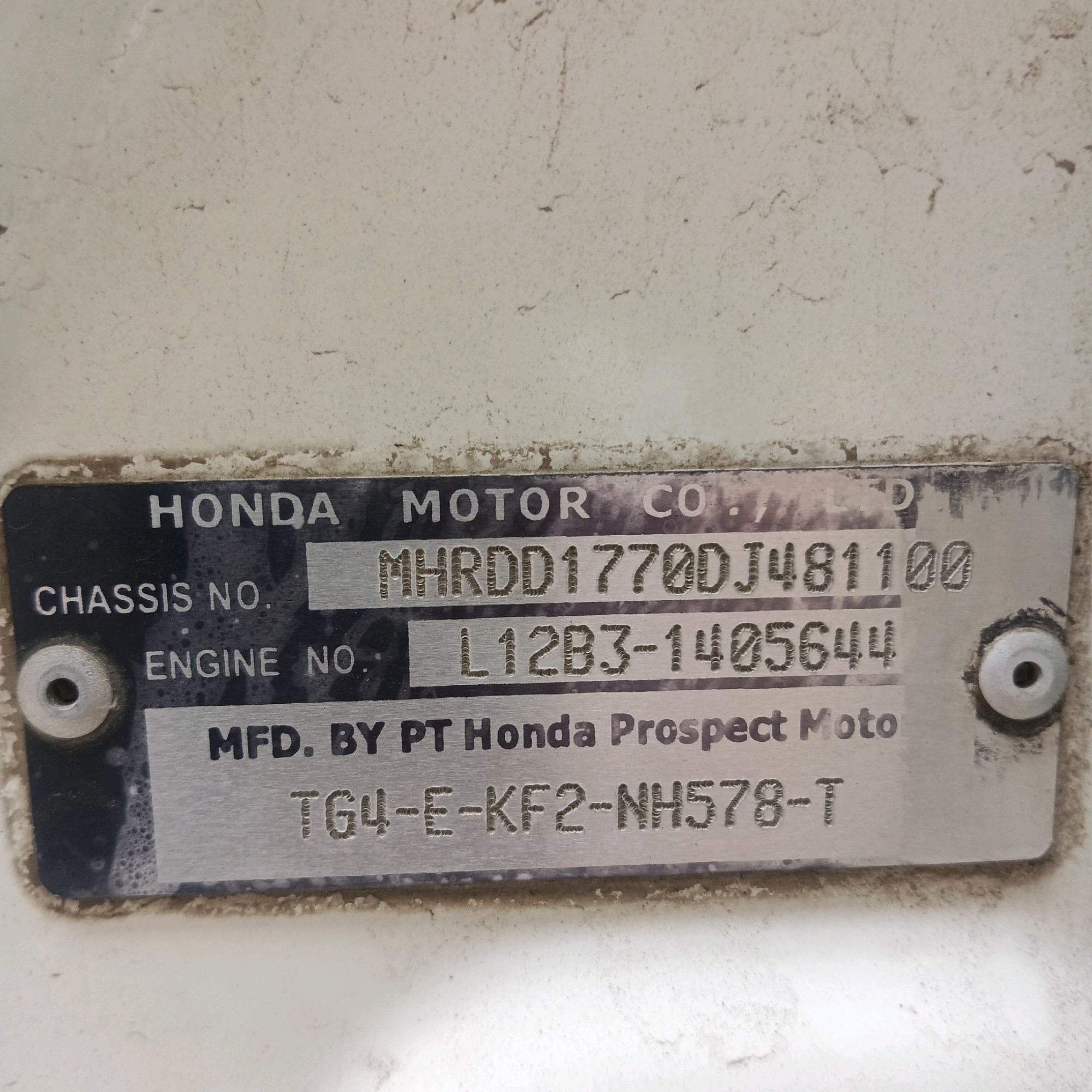 A rectangular metal plate is affixed to a light-colored surface, likely a vehicle part. The plate is engraved with text and numbers. At the top, "HONDA MOTOR CO." is visible. Below this, "CHASSIS NO." is followed by "MHRDD1770DJ481100". Next to "ENGINE NO." is "L12B3-1405644". The bottom line reads "MFD. BY PT Honda Prospect Moto" with a serial number below it: "TG4-E-KF2-NH578-T". The plate has two circular metallic fasteners, one on the left and another on the right. The surface appears dusty and weathered. The scene is indoors, with no discernible city or location information present in the image.