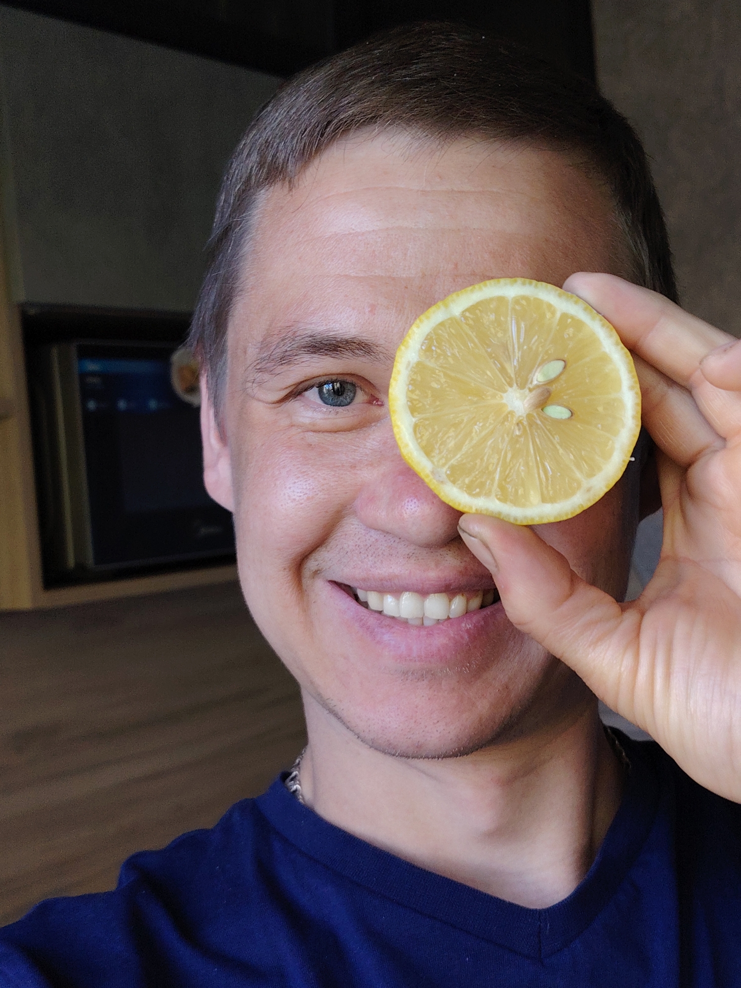 A smiling man is holding a lemon slice in front of his right eye. He has short brown hair and blue eyes. He is wearing a dark blue V-neck shirt. A stainless steel microwave with a black frame is visible in the background. The location is in a residential interior in the city of Chaikovsky, Russia.