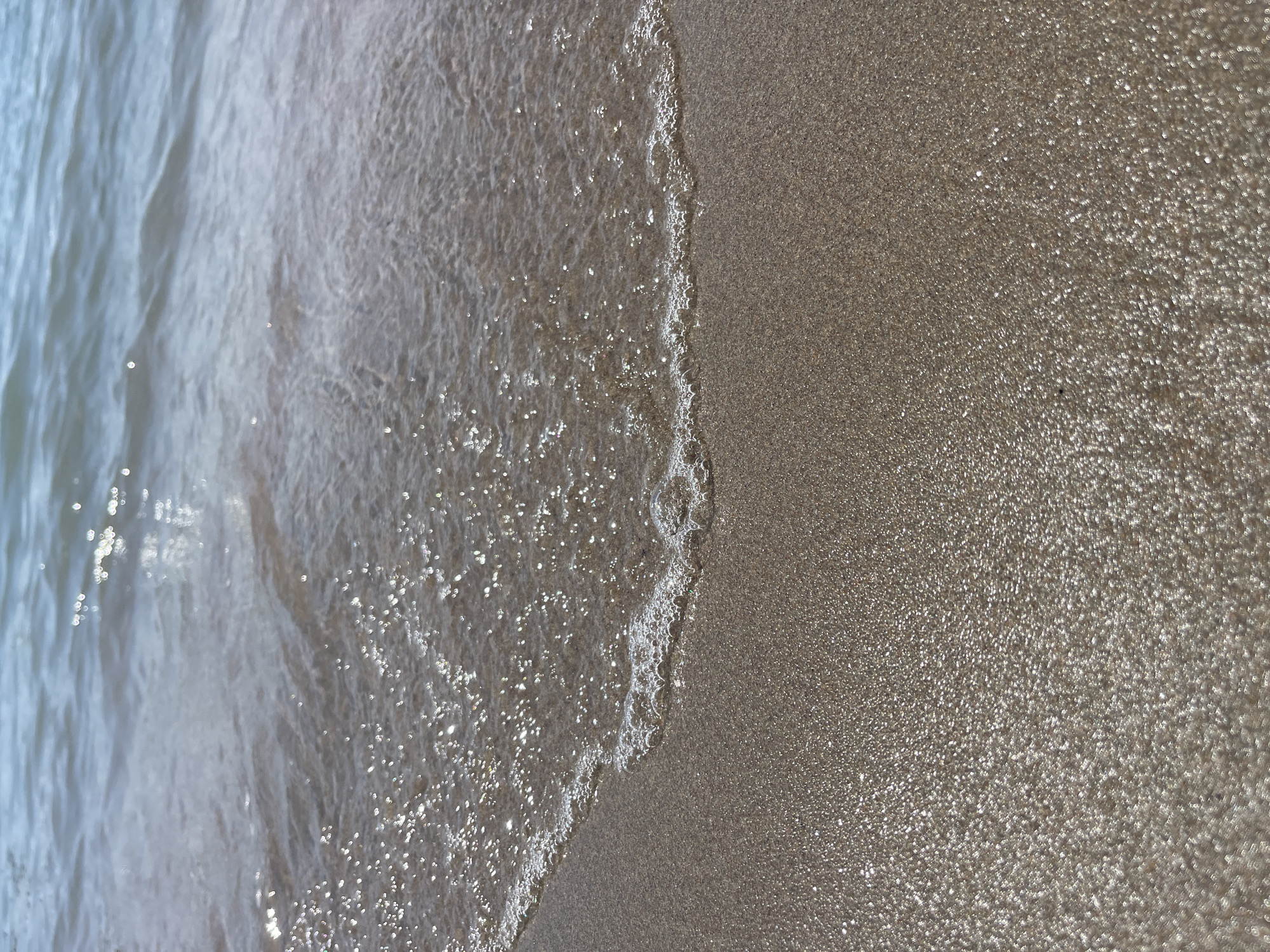 The image is a close-up, eye-level shot of a sandy beach with gentle waves lapping at the shore. The sand is a light brown color and appears wet and glistening in the sunlight, with small particles of shell or other debris visible. The water is clear and blue, with a soft ripple pattern and bright highlights from the sun reflecting off its surface. A thin line of white foam and bubbles marks the edge where the water meets the sand. The scene evokes a peaceful and serene beach environment. The location is not specified in Ornek, Kyrgyzstan.