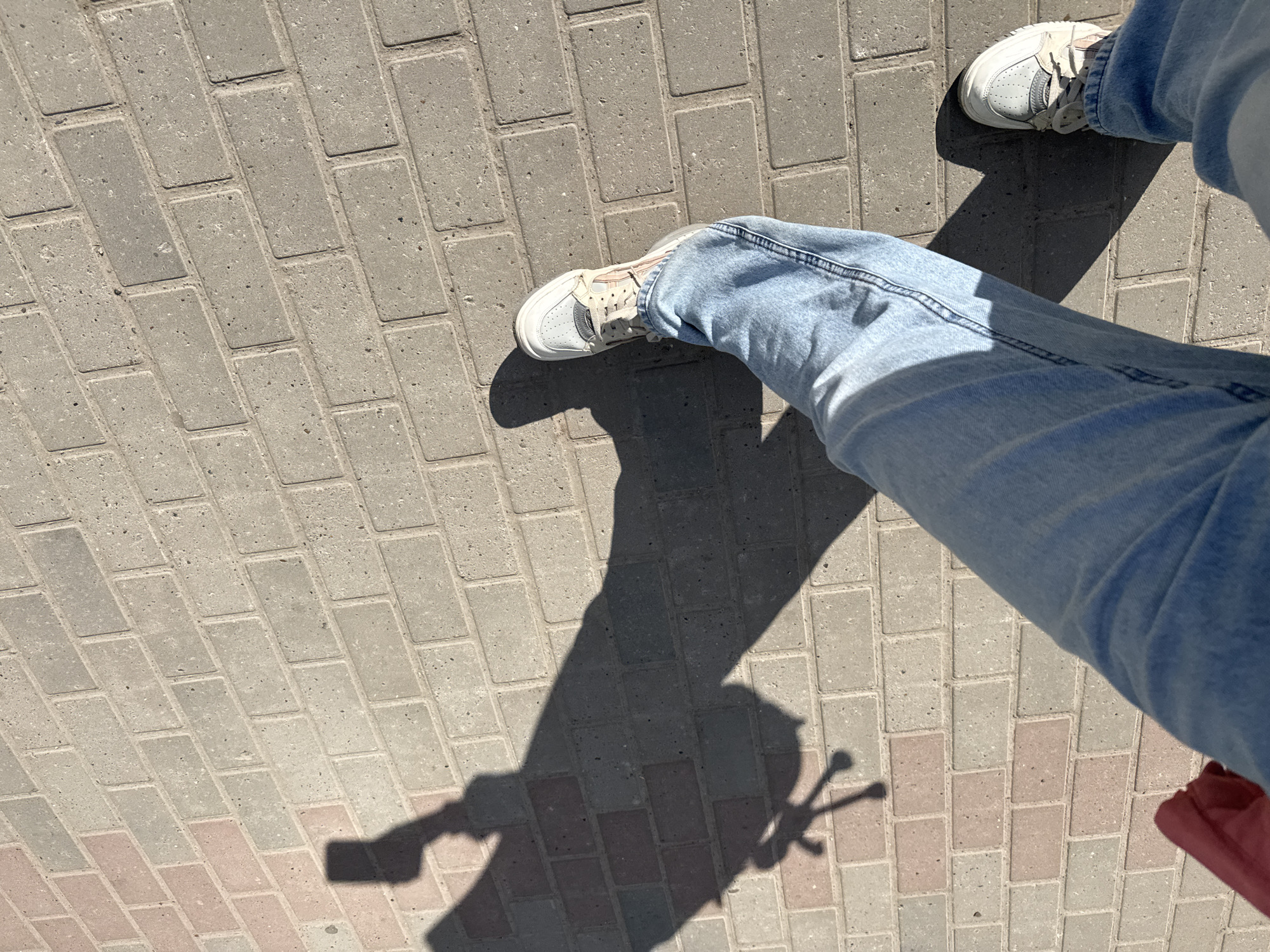 The image shows a person's legs and shadow on a brick walkway. The person is wearing light blue jeans and white sneakers. The walkway is made of gray and pink bricks. The shadow suggests the person is holding an object with a handle. The location is likely outdoors in Chaikovsky, Russia, based on the information provided.