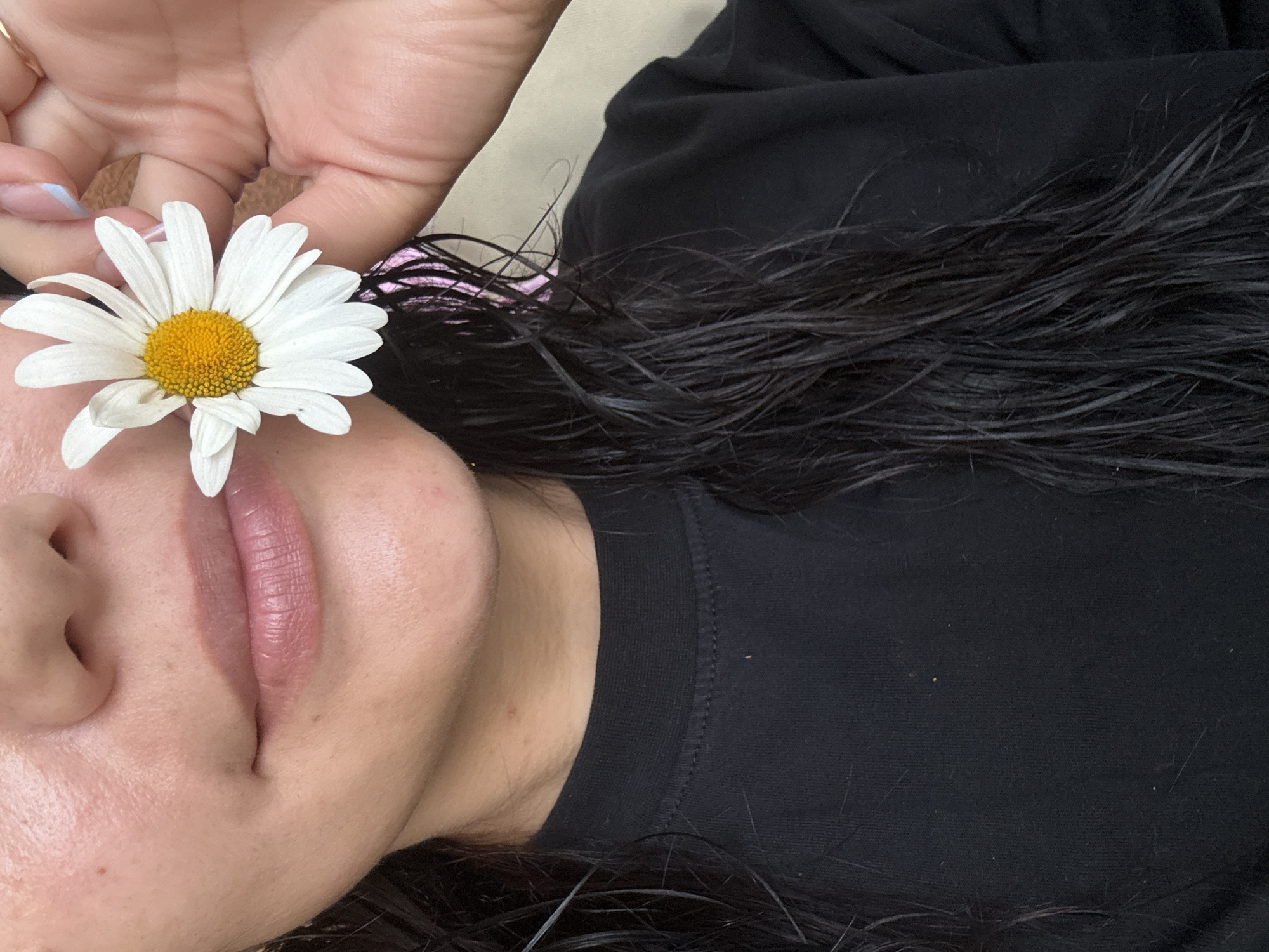 The image shows a close-up of a person's face, specifically their mouth and nose area, and a portion of their chest and neck. The person is holding a white daisy with a yellow center up to their face, near their nose. Their lips are lightly colored with lipstick. The person has dark, wet hair falling around their shoulders. They are wearing a black crew-neck t-shirt. The background is a neutral, beige color. There is no discernible information in the image that could indicate a location in Saint Petersburg, Russia.