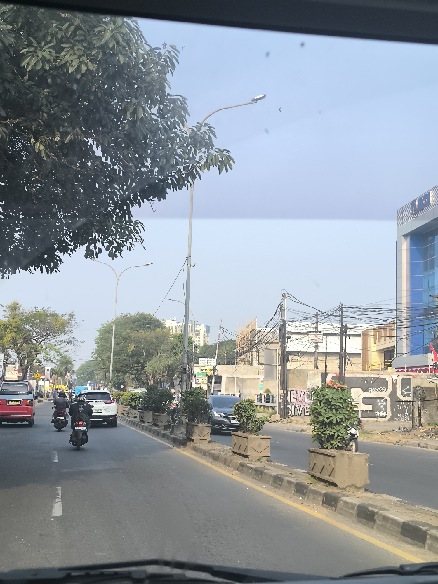 The image captures a street scene in Tangerang, Indonesia, likely during the daytime. The perspective is from inside a vehicle, looking forward through the windshield. The road is paved and appears to be multi-lane, with a yellow line marking the edge and a dashed white line dividing lanes. Several vehicles are visible on the road, including a red car, a white SUV, and at least two motorcycles. The motorcycles are being ridden by individuals wearing helmets and jackets.
The left side of the road features a sidewalk with planters containing green shrubs. Beyond the sidewalk, there are trees and some buildings. A large tree with dark green foliage is prominent in the foreground on the left. Streetlights are also present.
The right side of the road shows a sidewalk with more planters, some with flowering plants. Beyond the sidewalk, there is a building with a modern facade, featuring blue glass windows and a gray upper section with lettering that is partially obscured but appears to include "AMG". Further down the street, there is ongoing construction with scaffolding and concrete structures. Overhead, a dense network of electrical wires is visible, a common sight in urban Indonesian environments. Graffiti can be observed on some of the lower walls. The sky is clear and light blue, suggesting fair weather.