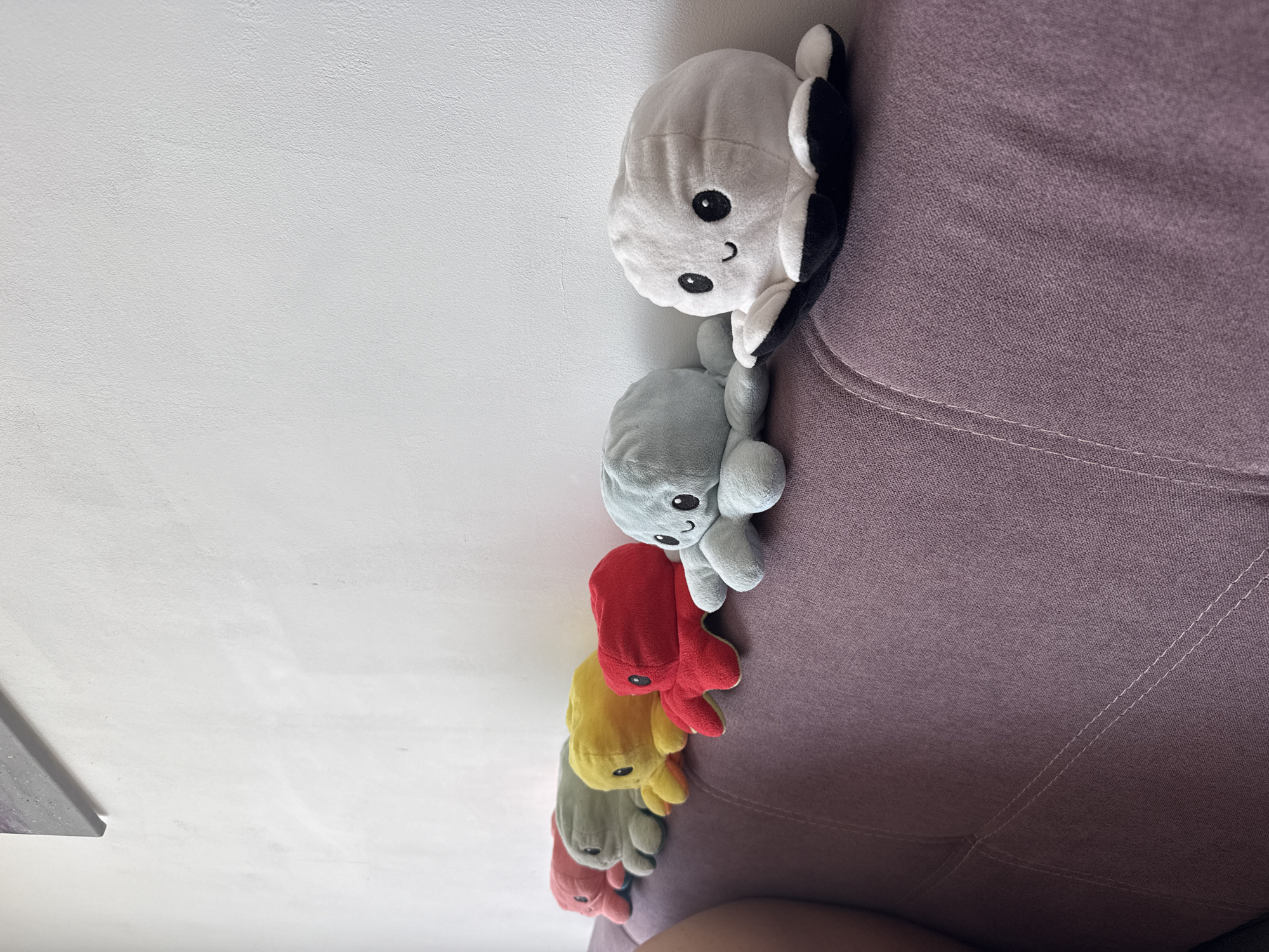 The image shows a row of reversible octopus plush toys arranged on a lavender couch against a white wall. From top to bottom, the colors are white and black, gray, red, yellow, green, and pink. The photo was taken indoors, possibly in a home. The location is not identifiable as Plovdiv, Bulgaria.