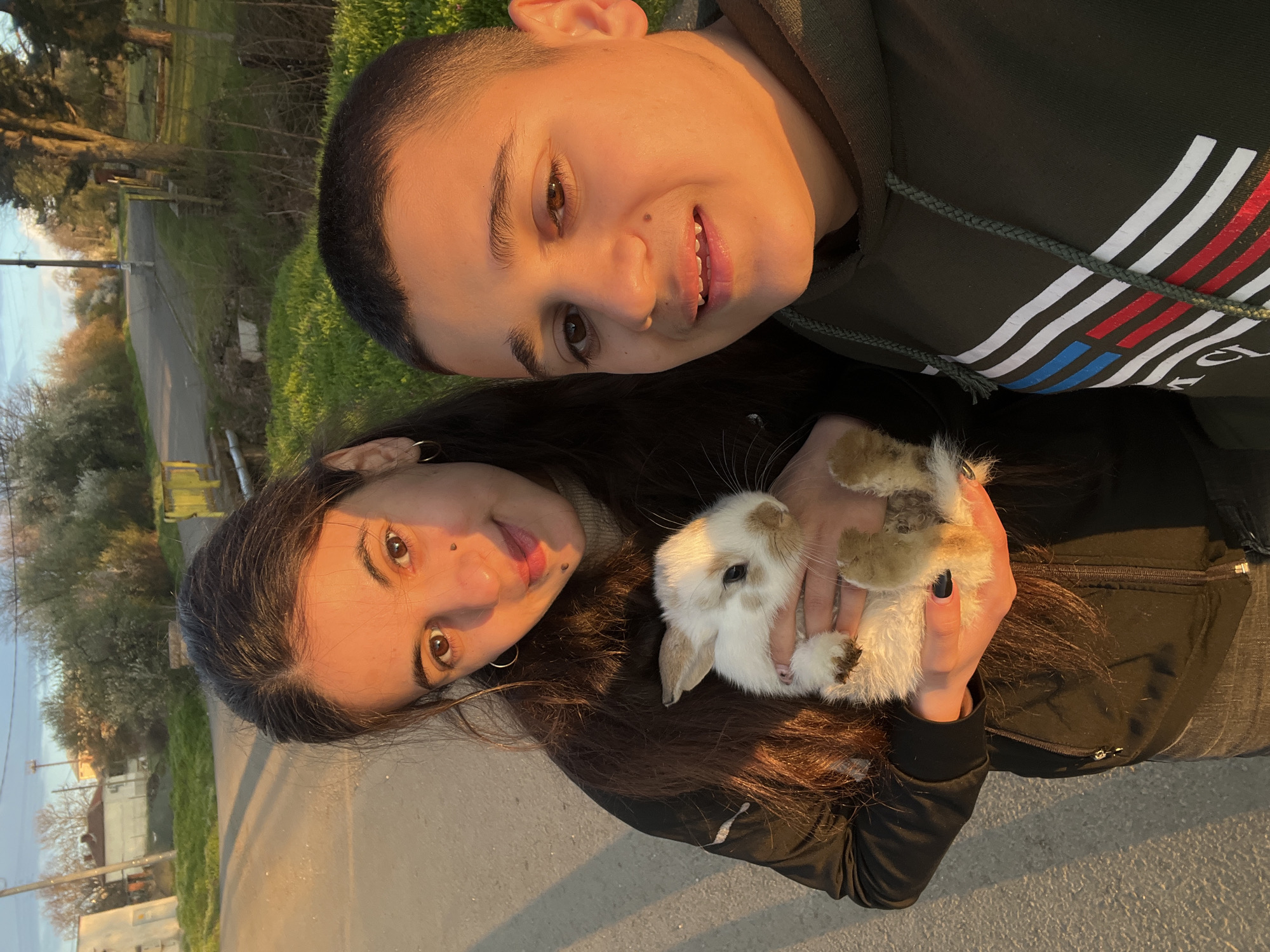 Two individuals and a rabbit are in the frame. One person with short hair is wearing a hoodie with stripes and is smiling. The other person, with long dark hair, is holding a rabbit. They are on a road with greenery on either side. A building and trees are visible in the background. The image appears to have been taken in natural light, possibly during sunset. There are no other indicators of a specific location in Lozarevo, Bulgaria, beyond the general landscape.