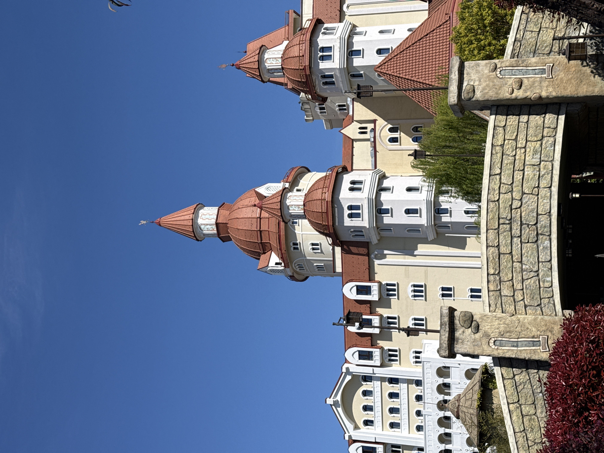 The image presents a grand and ornate building, possibly a hotel or resort, featuring a distinctive architectural style reminiscent of a fairytale castle, set against a vibrant clear blue sky. The multi-storied structure boasts a cream-colored facade with intricate white detailing around numerous windows, some arched and some rectangular. Dominant features include multiple cylindrical towers and turrets, all crowned with reddish-brown tiled roofs. One prominent turret towards the left is topped with a pointed spire and a small weather vane.
On the right side of the image, heavy, textured stone-like architectural elements are visible, suggesting a bridge, archway, or decorative wall, complete with integrated stones and decorative insets. Green foliage from trees and bushes can be seen surrounding parts of the building, and a striking deep red/maroon bush occupies the lower right corner.
The photograph captures the scene on a bright and sunny day, indicated by the uniformly clear, cloudless blue sky and the crisp lighting. There is no discernible activity or specific event taking place, and no people are visible. The image focuses on the architectural beauty and the serene setting. No text is visible within the frame. Given the location context of Sirius, Russia, this building likely serves as a themed tourist destination or a luxury accommodation.
