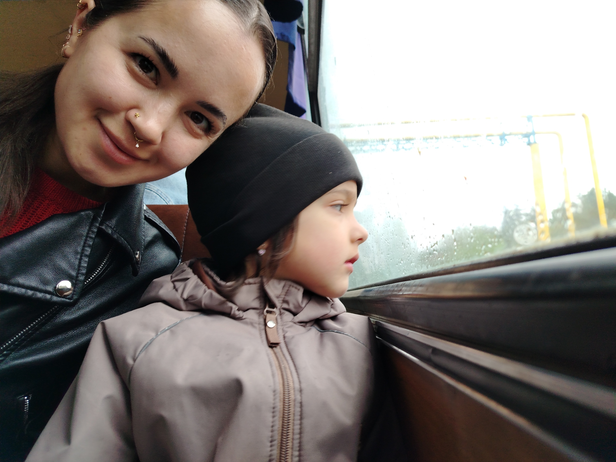 A woman and a child are sitting close together, likely on public transport, judging by the window. The woman is on the left, with dark hair, a nose piercing, and a black leather jacket. She is smiling slightly at the camera. The child, wearing a black beanie and a beige/grey hooded jacket, is looking out the window. The window appears to be wet, possibly due to rain. The background shows blurred scenery, and what appears to be overhead electrical lines. The location might be Noviy, Russia, as indicated in the instructions.