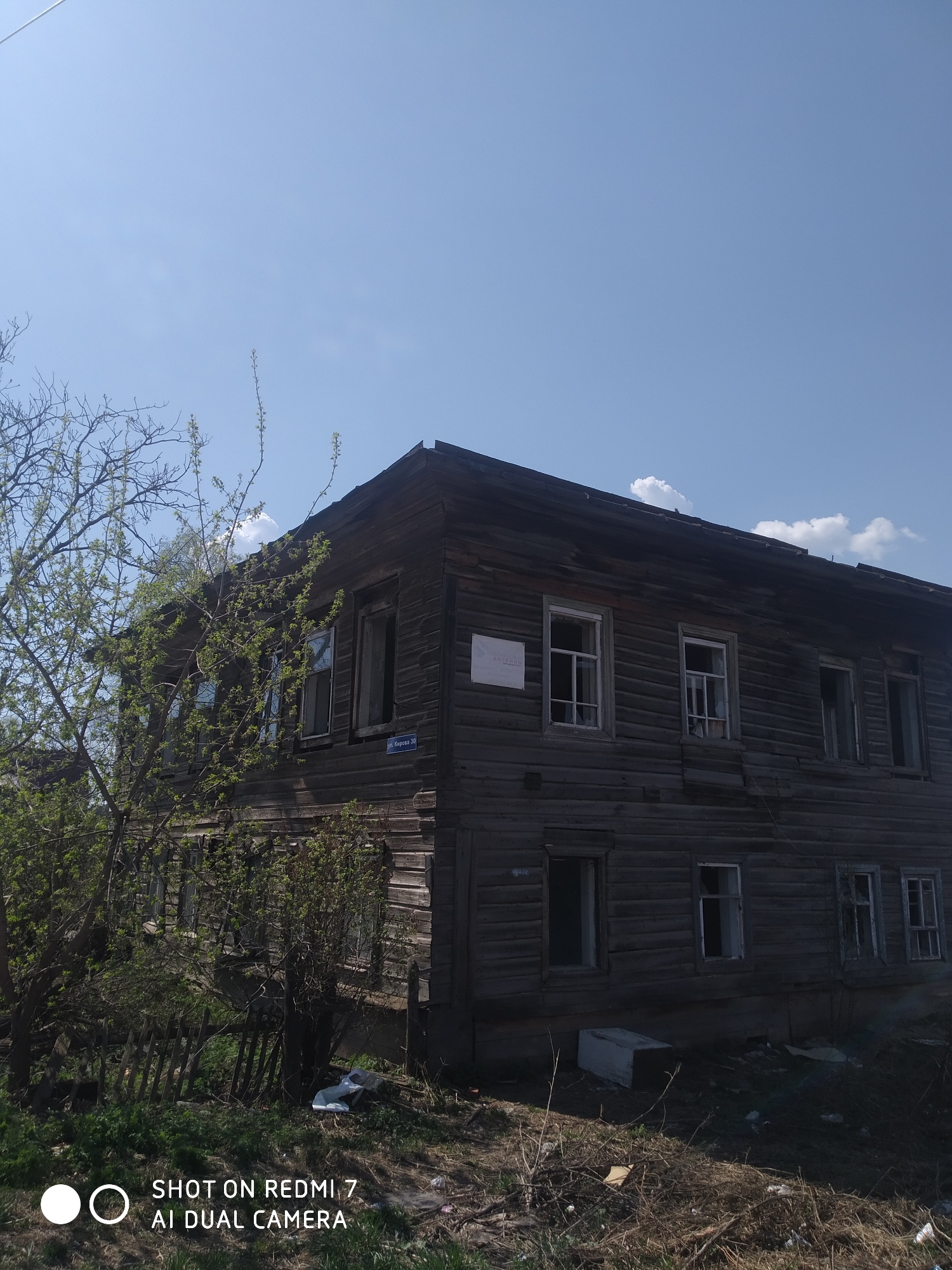 The image shows a dilapidated two-story wooden building under a blue sky with scattered clouds. The building has multiple windows, some boarded up or broken. A street sign reading "ул. Кирова 30" (Kirov street 30) is visible on the building's corner. A small, leafless tree is in the foreground, along with some debris and overgrown vegetation. The photo was taken with a Redmi 7 AI Dual Camera. The location is Фоки, Russia, based on the street name.
