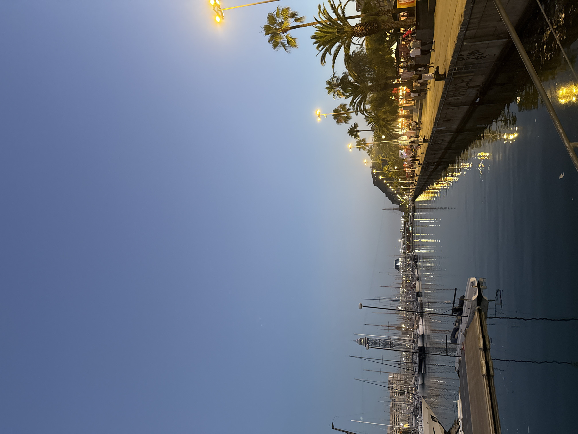This image captures a twilight scene in the Port Olímpic area of Barcelona, Spain. The view looks across a calm marina filled with numerous sailboats and a few larger yachts, their masts creating a dense forest of vertical lines. A modern-looking tower, possibly a lighthouse or observation point, is visible in the distance among the boats.
To the right of the marina, a wide, paved promenade runs along the waterfront. This promenade is illuminated by rows of tall lampposts with bright yellow lights, casting reflections on the water and the pathway. Palm trees line the promenade, adding to the Mediterranean ambiance. Several people are strolling along the promenade, some in small groups, enjoying the evening. A few structures, potentially shops or restaurants, are situated behind the palm trees. The sky is a clear, deep blue, transitioning from the fading daylight. The overall atmosphere is peaceful and suggests a pleasant evening at a popular waterfront location.