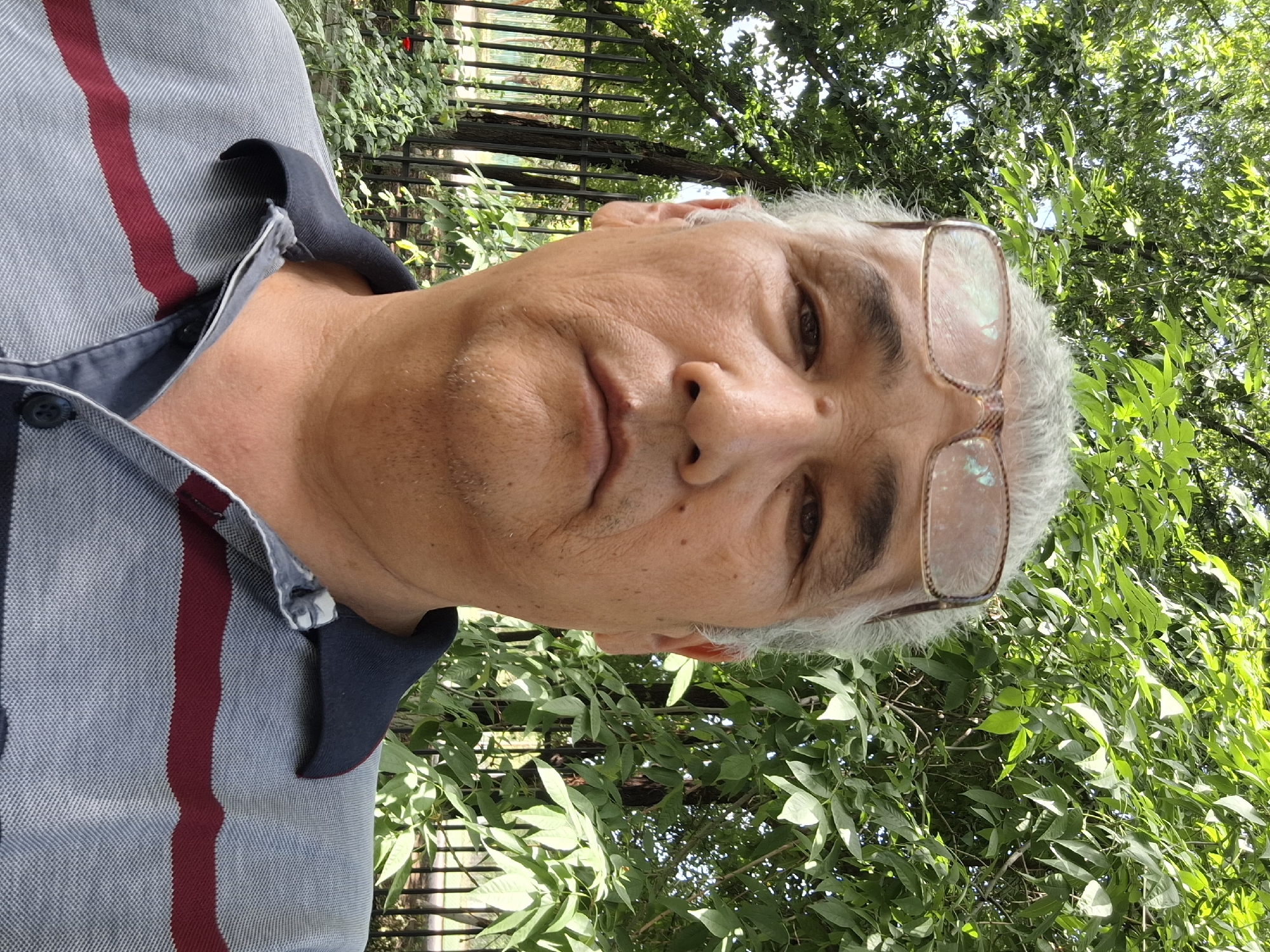 A man with graying hair and glasses is pictured outdoors, facing the camera. He is wearing a collared shirt with horizontal stripes. The background consists of lush green trees and foliage, suggesting a park or natural area. The lighting indicates it is daytime. There is a black metal fence visible behind him. The scene appears to be in Almaty, Kazakhstan, given the reference to the city.