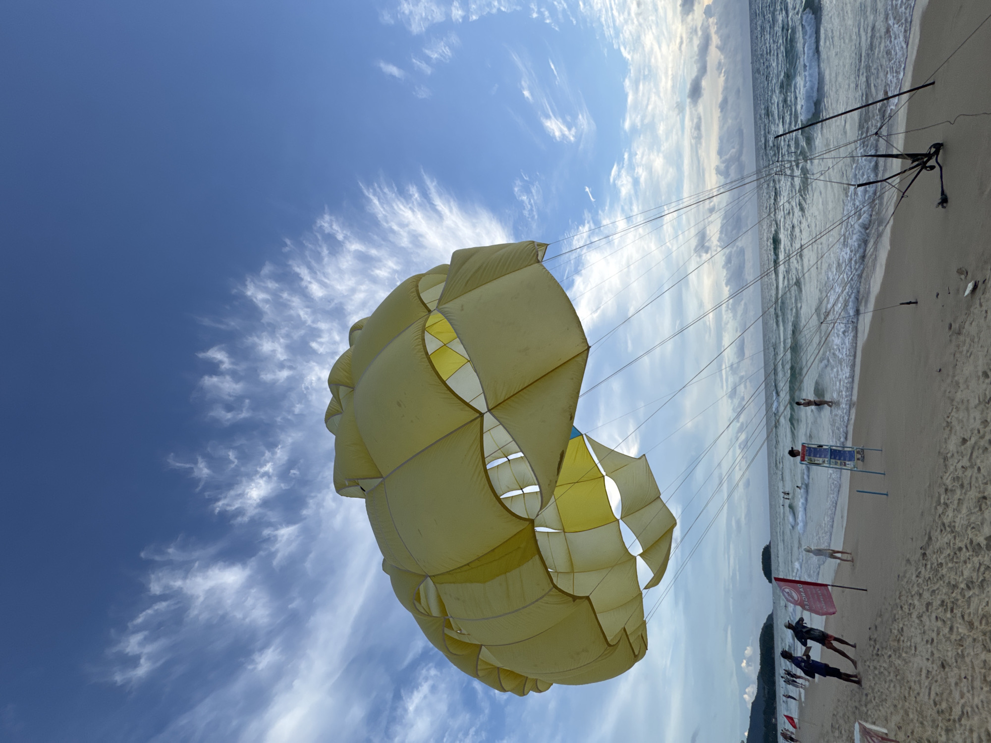 A bright yellow parachute, presumably for parasailing, fills the majority of the frame, angled upwards towards a vibrant blue sky dotted with wispy white clouds. The parachute is intricately constructed with a diamond-patterned design in some sections. Below the parachute, a sandy beach stretches towards a calm ocean. The water exhibits gentle waves crashing onto the shore. Several people are visible on the beach and in the shallow water, some appearing to be enjoying the beach or preparing for activities. A dark metal structure, possibly a stand for the parasailing equipment, is set up on the sand near the water's edge, with numerous lines extending upwards towards the parachute. The scene suggests a sunny day, possibly late afternoon given the quality of light. The location is identified as Karon, Thailand. There is a red flag planted in the sand with white text that is not clearly legible. A signpost with multiple languages is also visible on the beach.