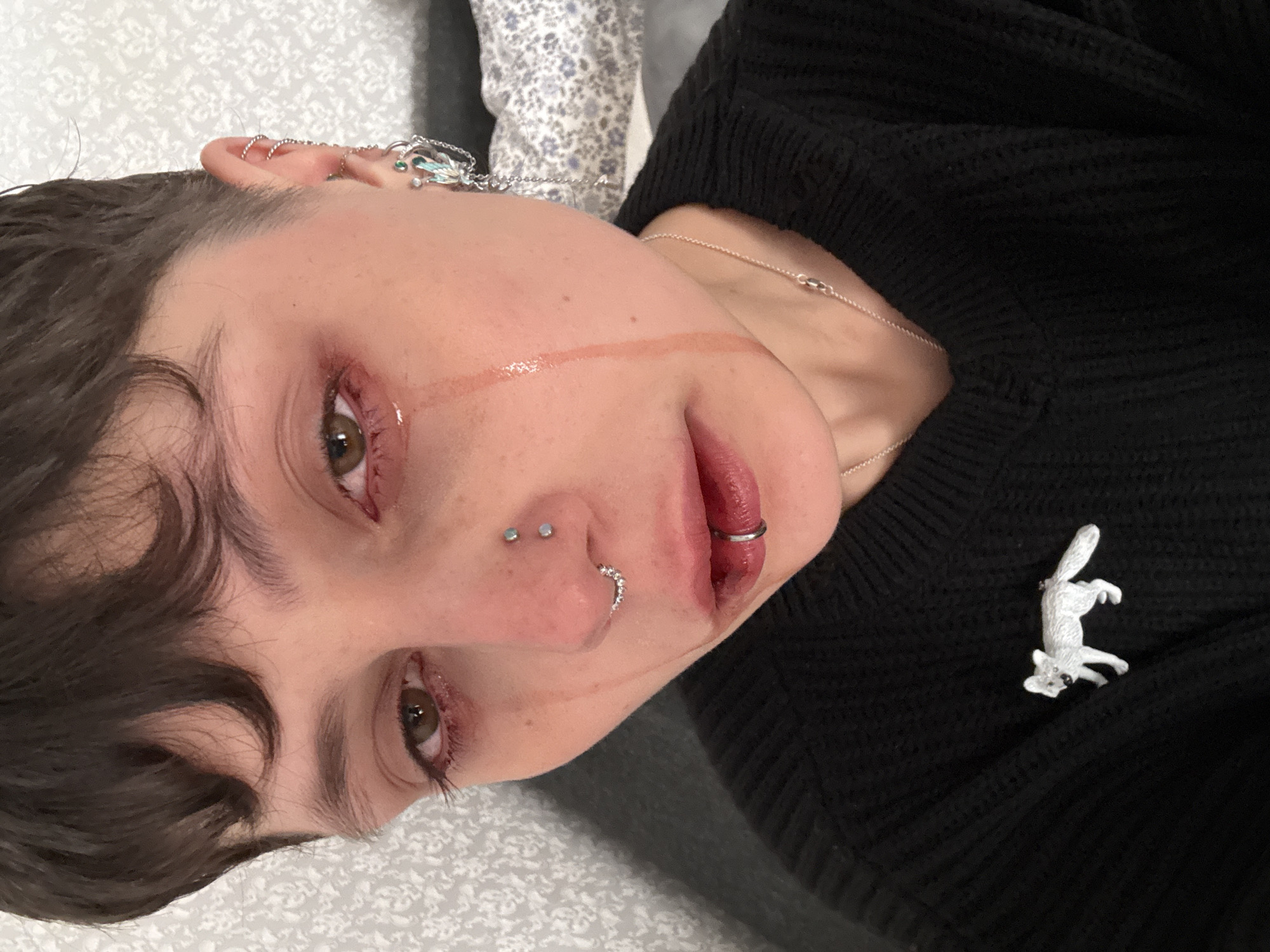 A close-up, portrait-style photograph depicts a person with fair skin, short dark hair with a slight wave on the left side of the frame, and light brown eyes with visible redness around the lids. The person has multiple piercings on the ear and nose, including a nostril piercing, a septum piercing, and several cartilage piercings. They are wearing a silver lip ring and a silver necklace. A streak of a clear, glossy substance is visible from the corner of the left eye down the cheek.
The person is wearing a black, knitted sweater adorned with a small, white fox-shaped pin on the right side. The background consists of a light-colored surface with a subtle pattern, possibly a bedspread or couch.
There is no information to suggest a location in Saint Petersburg, Russia.