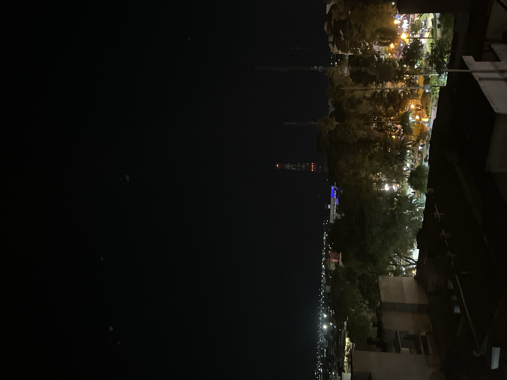 The image shows a nighttime view of San Fernando del Valle de Catamarca, Argentina. In the foreground, parts of buildings are visible, including walls and roofs. Beyond these, the city stretches out, illuminated by streetlights and building lights. Tall communication towers are prominent in the distance. A line of trees runs through the city, and park lights are visible among the trees. In the dark sky, the faint outline of distant hills can be seen.