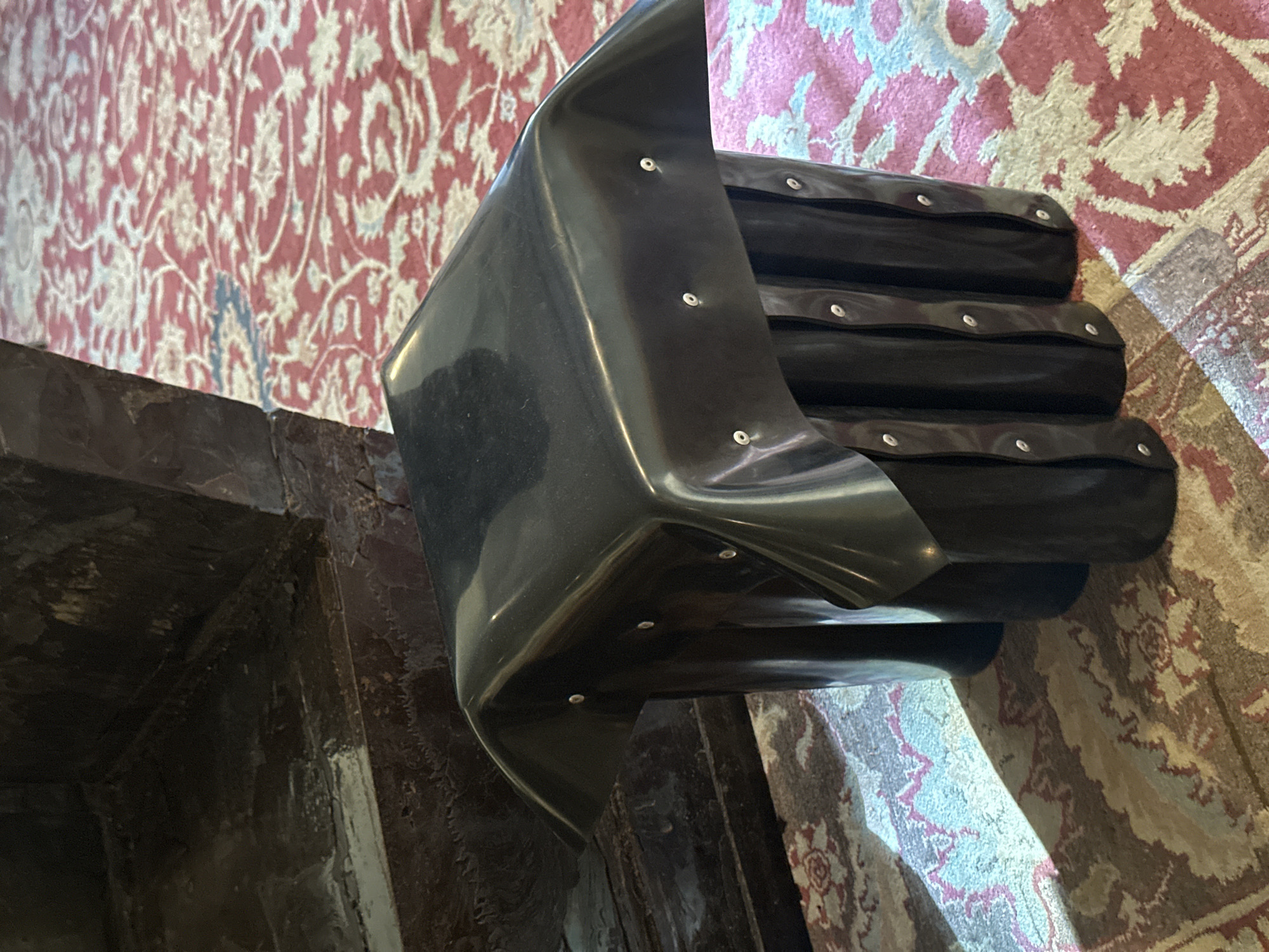 A low-angle, close-up photograph depicts a black, geometric chair resting on a patterned rug. The chair features a glossy, curved back and a tiered seat, each tier secured with metallic rivets. The rug exhibits a complex pattern of red, beige, and blue hues. To the left of the chair, a dark, textured structure appears to be a portion of a building or other architectural element. The location is presumed to be in the city of London, United Kingdom.