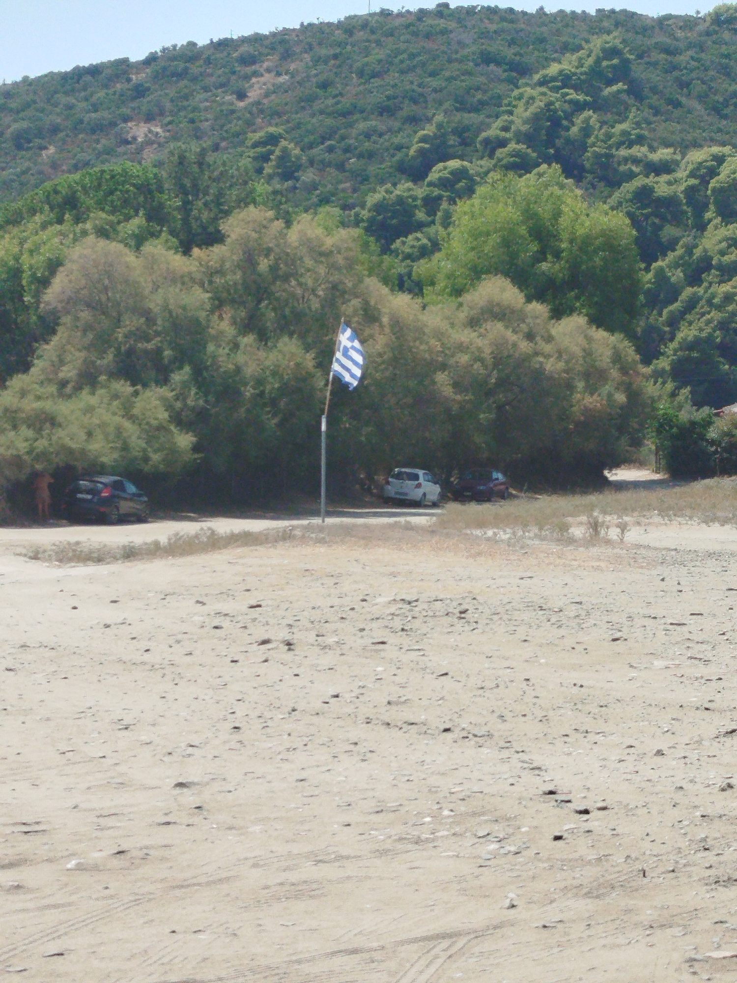 The image depicts a sparsely populated beach area near Sithonia Municipal Unit, Greece. The foreground is dominated by sandy terrain with scattered pebbles and tire tracks. In the midground, a Greek flag on a pole stands prominently, positioned between a line of dense green trees and a more sparse, lighter green shrubbery. Several cars are parked in the shade of the trees, including a dark colored sedan on the left and a white and a dark red car further to the right. The background consists of a densely vegetated hillside, characterized by a mix of dark green trees and lighter green bushes, under a clear blue sky. One person is partially visible near the dark sedan, suggesting a low level of human activity. The scene conveys a sense of a quiet, possibly remote, coastal location.