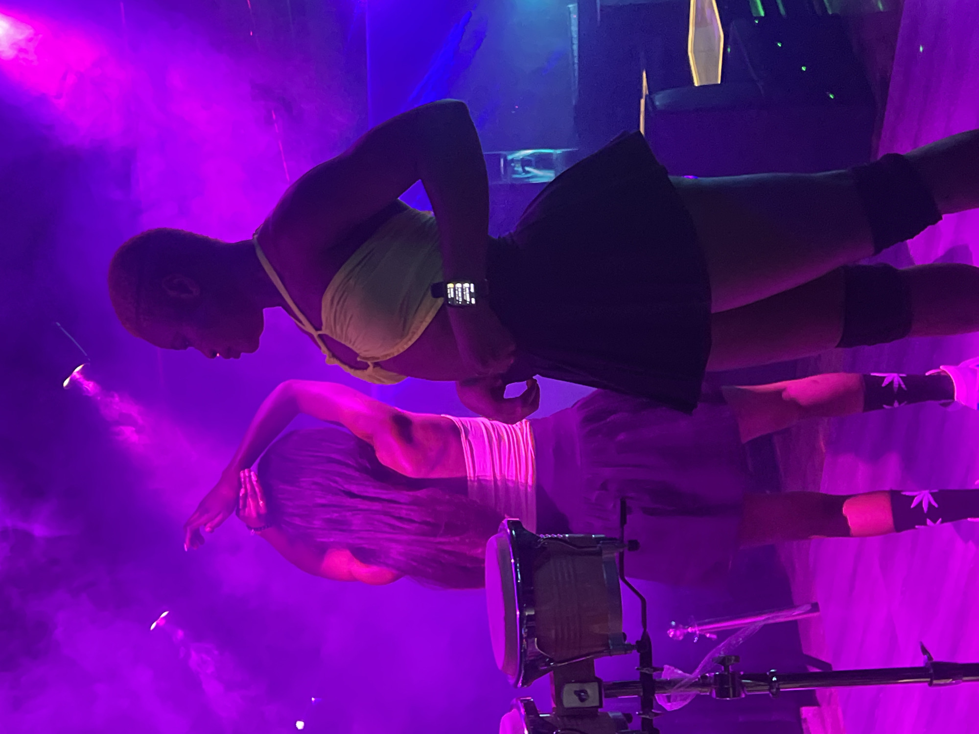In a dimly lit club in Howayi, Nigeria, two individuals are seen performing on stage. The stage is bathed in vibrant purple and blue lights, with smoke machines creating a hazy atmosphere. The subjects are dressed in performance attire. The person in the foreground is wearing a light yellow halter top and a short black skirt, with black knee-high socks adorned with white cannabis leaf patterns. They are facing away from the camera and appear to be in motion.
The second performer, positioned behind and slightly above the first, is wearing a white pleated skirt and a white top. Their hair is long and appears to be dark. They are seen from behind, with their arms gracefully extended.
In the lower right corner of the image, a drum set is partially visible, suggesting a musical performance is taking place. The overall mood of the image is energetic and dynamic, typical of a live performance or club event. The time of day is likely evening or night, given the indoor setting and the use of stage lighting. There are no visible signs of weather conditions. The specific location cues are limited to the context of a performance space. The visible text is on a watch worn by the foreground performer, showing a digital display.