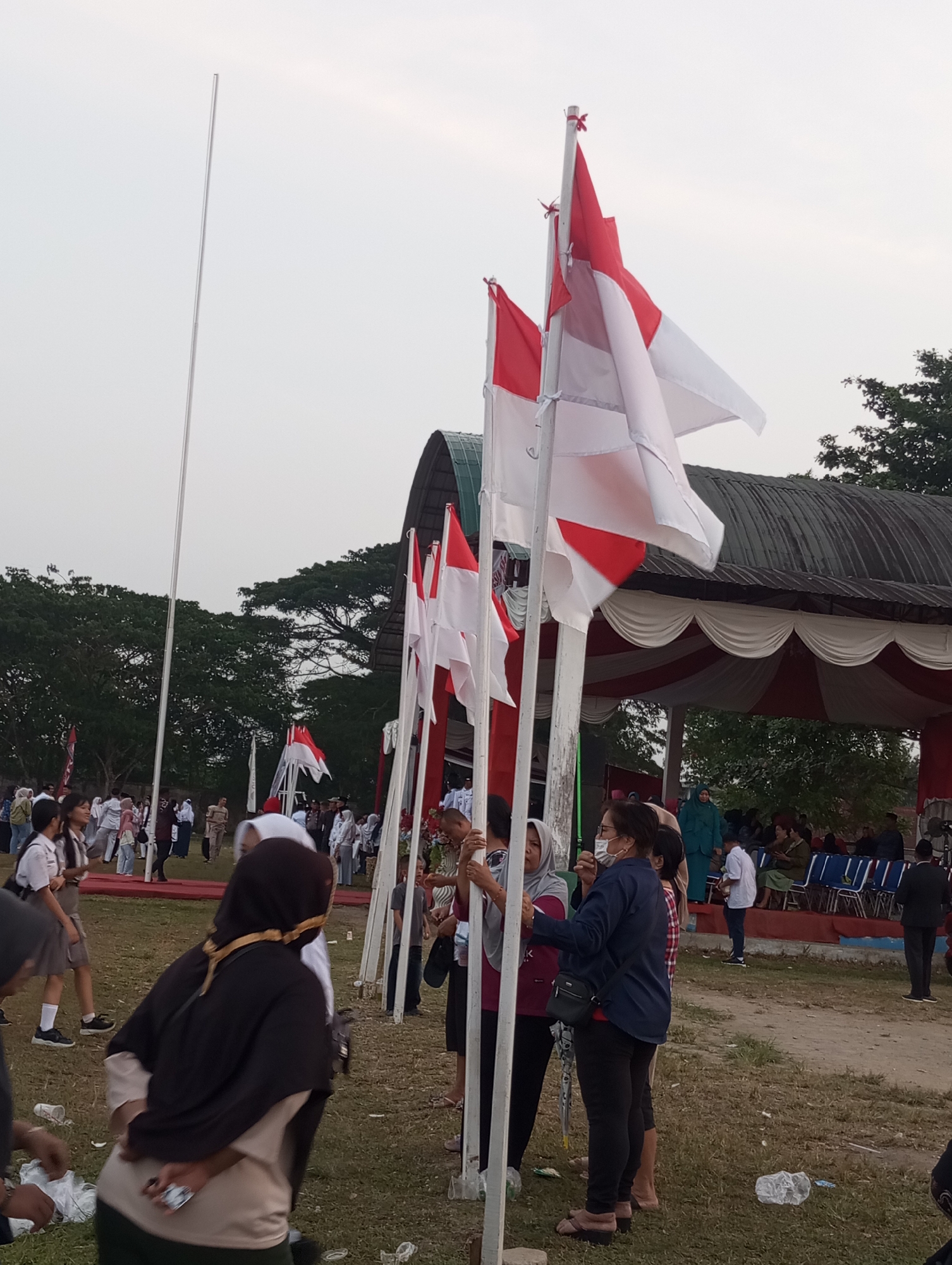In Candirejo, Indonesia, on a grassy field, an outdoor event is taking place. Several Indonesian flags, red and white, are prominently displayed on tall poles. A group of people, including children in school uniforms, are gathered, some observing the flags and others interacting with each other. In the background, a covered stage with red and white drapes suggests a performance or ceremony. The sky is overcast, and trees frame the scene. The overall atmosphere appears to be one of communal gathering and national pride.