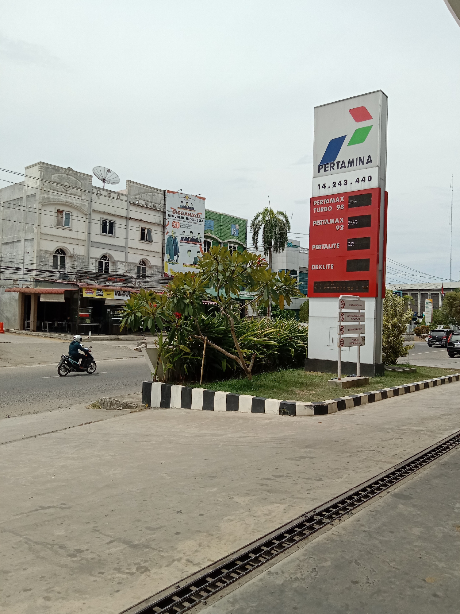 The image captures a street scene in Lhokseumawe, Indonesia, featuring a Pertamina gas station price sign. The sign displays prices for Pertamax Turbo 98, Pertamax 92, Pertalite, and Dexlite. Below the price display are signs in Indonesian prohibiting activities like smoking, using mobile phones, drawing graffiti, and igniting fires.
Adjacent to the gas station sign is a landscaped area with various plants and a curb painted with black and white stripes. The street view includes a building with a banner celebrating the Republic of Indonesia, a motorcycle rider, and other buildings in the background. The sky is overcast, suggesting a cloudy day. A drain runs along the bottom right of the frame.