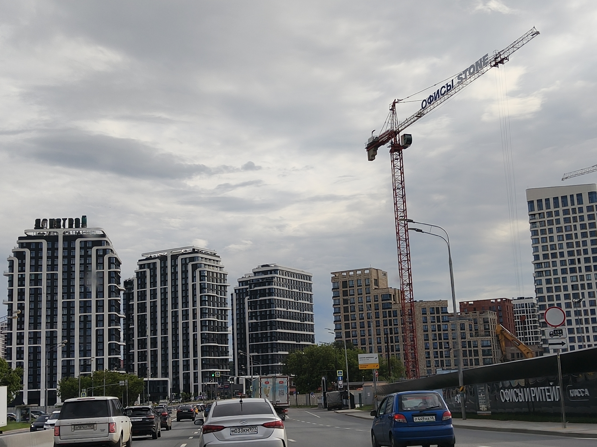 The image depicts a street scene in Moscow, Russia, featuring several modern high-rise buildings under construction. A large red construction crane dominates the sky, with "ОФИСЫ STONE" (Offices Stone) written on its boom. In the foreground, a busy multi-lane road is filled with various vehicles, including cars, SUVs, and a truck. The buildings exhibit contemporary architectural styles with dark facades and white accents. Some of the buildings appear completed, while others are still in various stages of construction. The sky is overcast with clouds. There are no people clearly visible in the image. The overall impression is one of urban development and ongoing construction projects in a city environment.