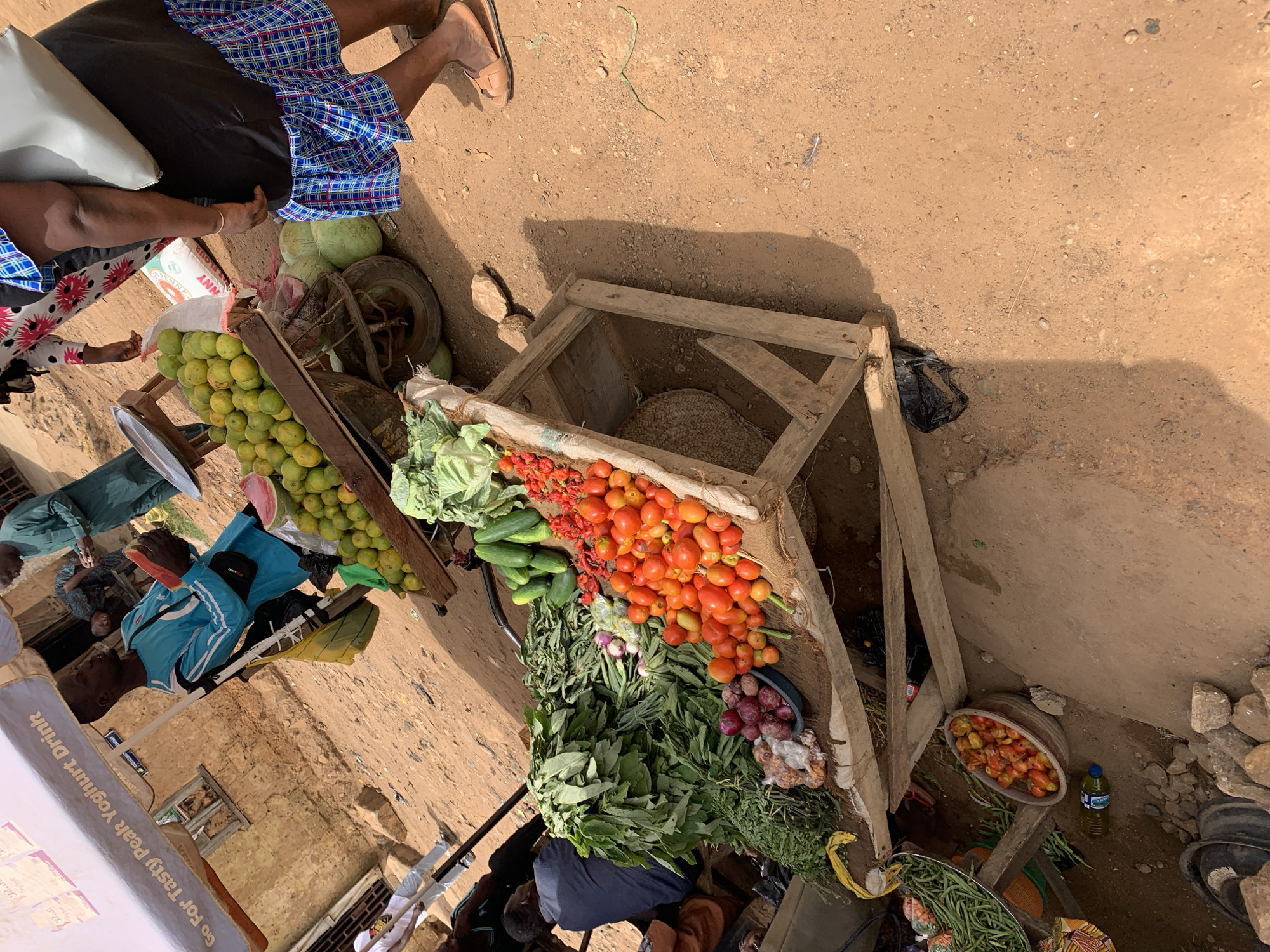 The image depicts a bustling marketplace scene in Rafin Par, Nigeria. From an elevated perspective, a vendor is selling a variety of fresh produce, including tomatoes, cucumbers, green leafy vegetables, onions, and mangoes, from a wooden cart and a wooden structure placed on the dusty ground. Some individuals are seen examining or purchasing the produce, while others are passing by. The background reveals a building constructed of mud bricks and a sign for a yoghurt drink.