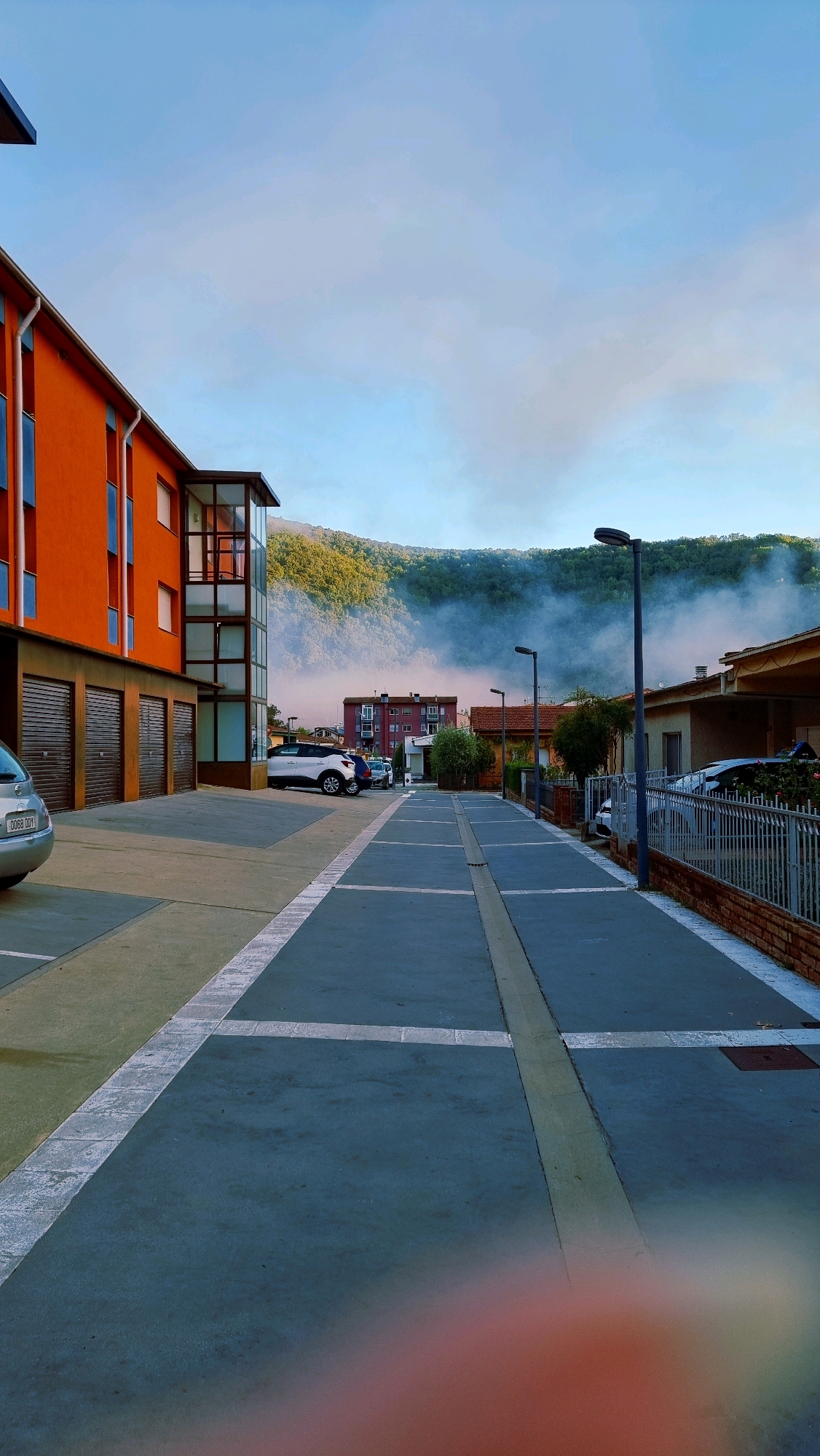 An outdoor, eye-level, full shot captures a modern urban street scene in Sant Joan Les Fonts, Spain. The morning light casts long shadows across the pavement, illuminating the scene with a soft glow. In the foreground, a gray asphalt parking lot is marked with white lines, dividing it into parking spaces. The lot is bordered by a sidewalk and a low brick wall topped with a metal fence. To the left, an orange building with blue-trimmed windows stands adjacent to a row of garage doors. Several cars are parked in the lot, including a silver car on the left, a white SUV, and another vehicle further down. In the background, a dense forest blankets a hillside, partially obscured by a wispy layer of fog or mist that hugs the treeline. The sky above is a clear, pale blue, with a few wisps of clouds. The overall impression is one of a quiet, orderly residential area at the edge of a natural landscape.