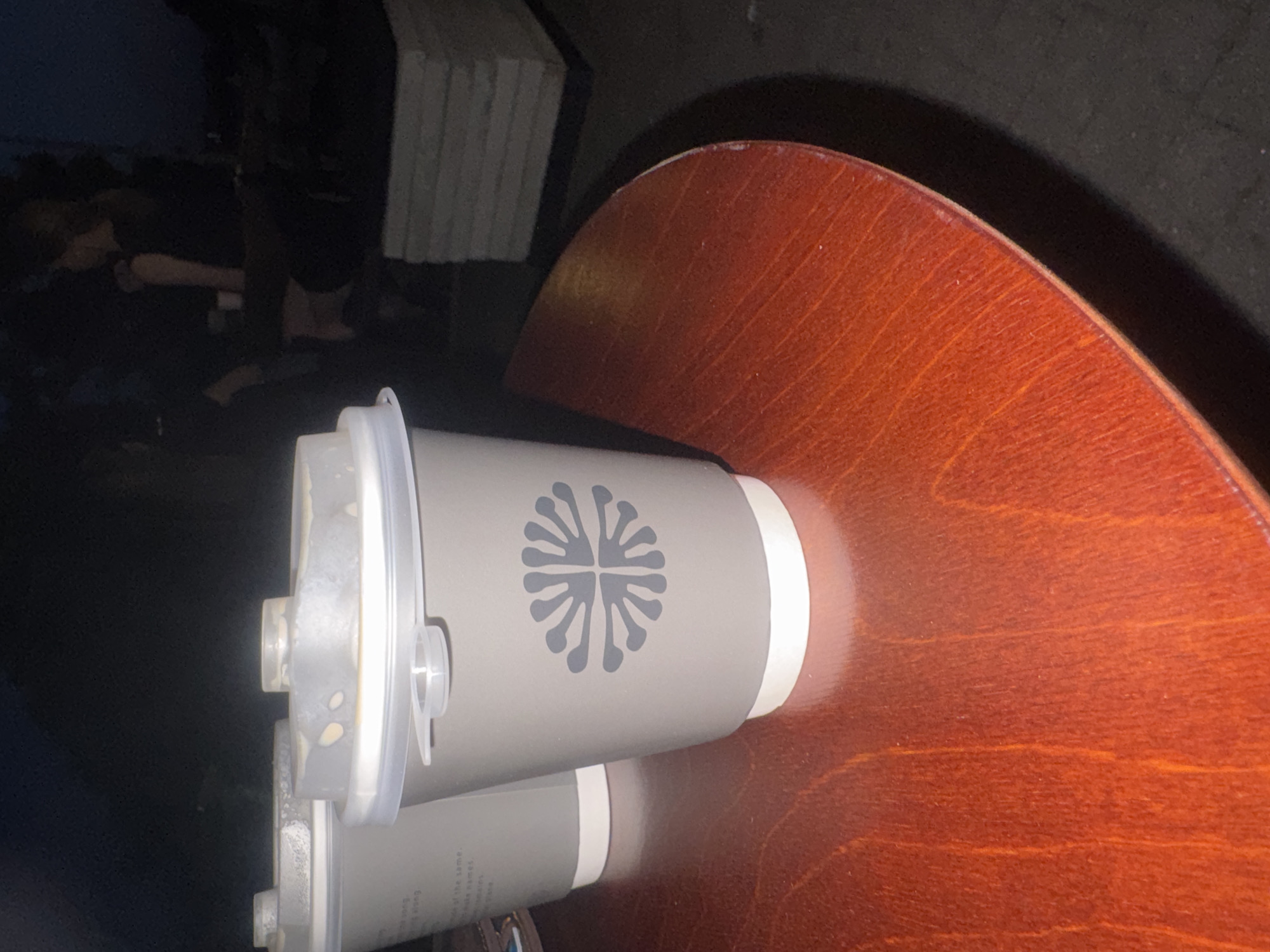 Two disposable coffee cups with grey sleeves and white lids are placed on a round, reddish-brown wooden table. The cup in the foreground has a dark grey stylized flower logo. The background is dark, with blurred figures of people and stacked objects visible. The scene is dimly lit, suggesting it is nighttime or an indoor setting with low lighting. The specific location in Krasnodar, Russia, cannot be determined from the visual information provided.