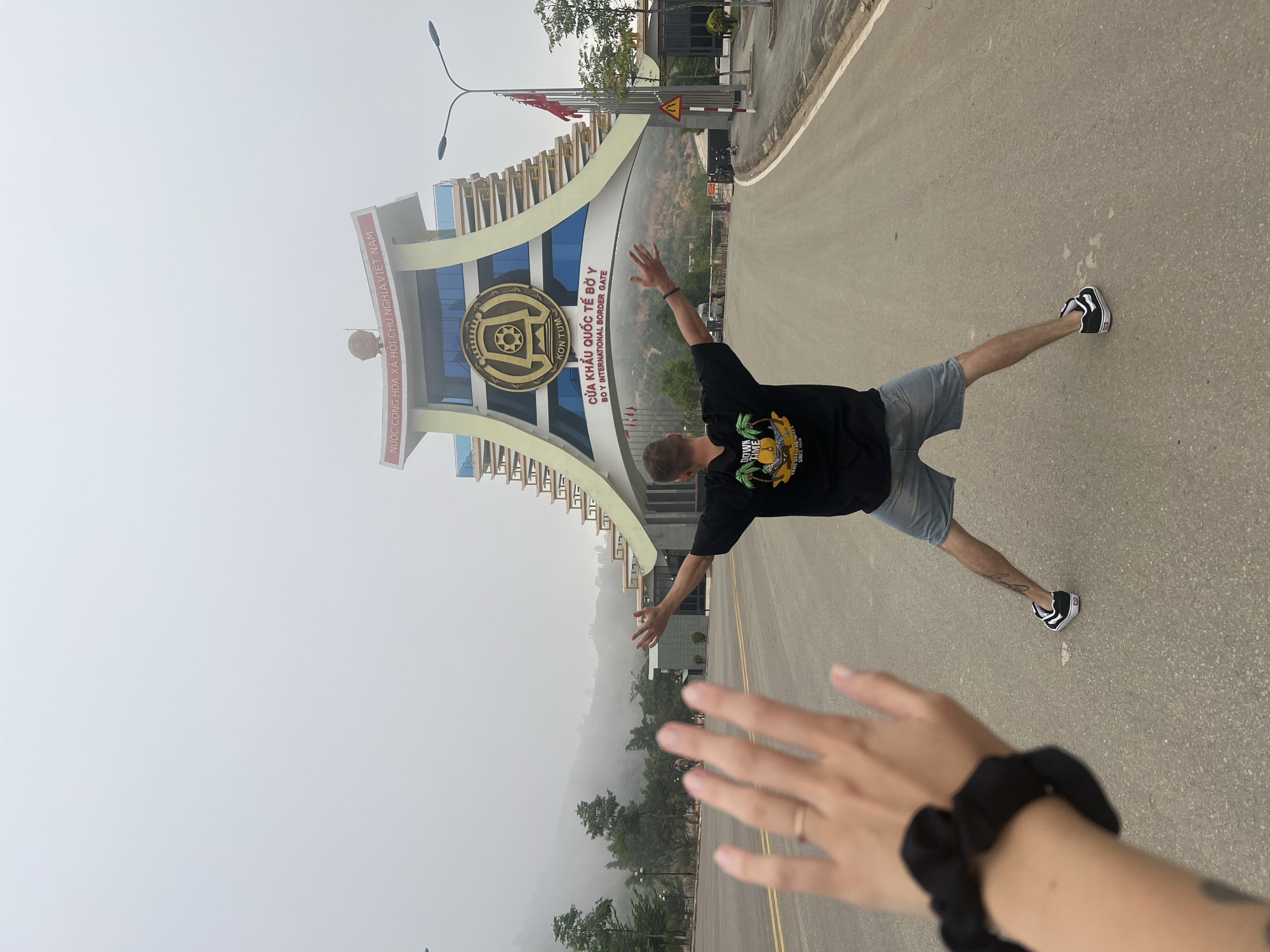 The image captures a person posing playfully in front of the Bờ Y International Border Gate in Kon Tum, Vietnam. A man, positioned on a paved road, extends his arms outwards and one leg sideways in a dynamic, almost leaping stance. He is dressed in a black t-shirt with a graphic reading "DOWN TIME" and "VANS OFF THE WALL SINCE 1966," light denim shorts, and black Vans sneakers with white soles.
The photograph is taken from a low angle, with the photographer's hand and forearm visible in the bottom right foreground, adorned with a black scrunchie and a ring. The background features the prominent border gate structure, characterized by its architectural curves, blue glass panels, and a large circular emblem in the center.
Visible text on the gate includes "NƯỚC CỘNG HÒA XÃ HỘI CHỦ NGHĨA VIỆT NAM" (Socialist Republic of Vietnam) and "CỬA KHẨU QUỐC TẾ BỜ Y" (Bờ Y International Border Gate), with "KON TUM" and "BO Y INTERNATIONAL BORDER GATE" also translated into English. Beyond the gate, the landscape is hazy and mountainous, with trees partially obscured by mist or fog, indicating an atmospheric day. The scene conveys a sense of excitement or triumph at reaching the border.