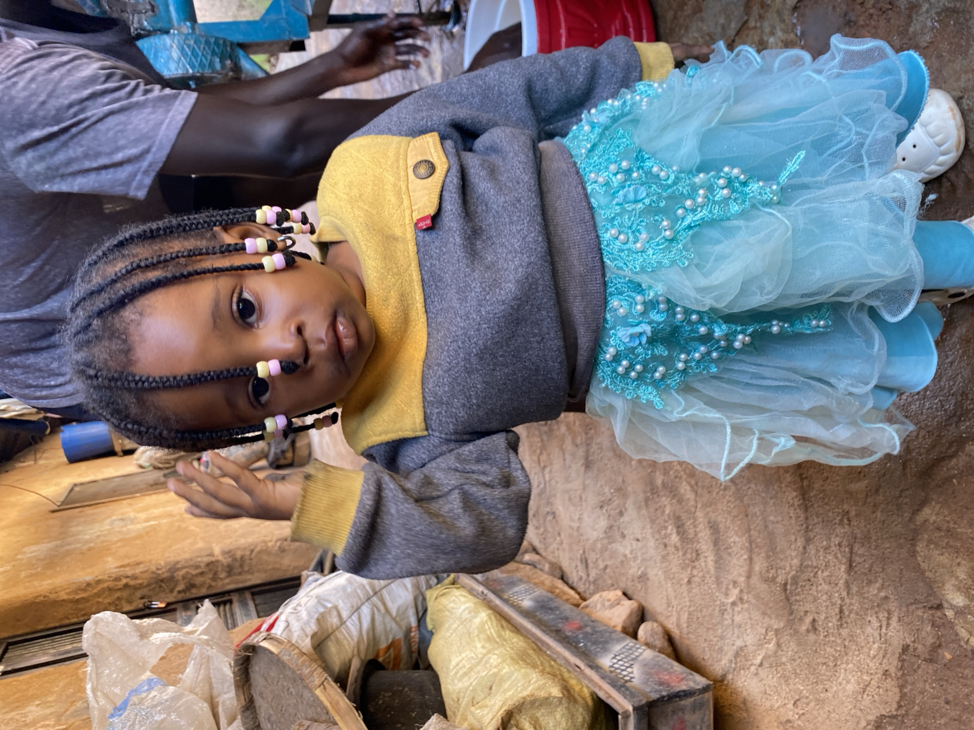 A young Black child with braided hair adorned with colorful beads is the central subject of this image. The child wears a gray sweatshirt with a yellow collar and chest detail, and a voluminous, layered turquoise skirt embellished with pearls and lace. The child's left hand is raised towards their head. The background suggests an outdoor setting in a seemingly less developed area, with a textured, earthy ground. To the left, there are stacked bags, one a large, light-colored woven bag, and a wooden crate containing a yellow-wrapped object. In the background, a portion of a building with a rough, ochre-colored wall is visible, along with a blue barrel. To the right, an adult's torso wearing a gray t-shirt is partially visible, and a red bucket is present. The scene appears to be a candid shot, capturing the child in a moment of interaction or pose.
The image depicts a scene likely in Jos, Nigeria, characterized by the outdoor environment with elements like the earthy ground, textured walls, and the presence of stacked materials often seen in urban or semi-urban settings in the region. The presence of the adult figure and the overall context suggest a domestic or community space. The child's attire, particularly the elaborate skirt, might indicate a special occasion or a display of local craftsmanship. The lighting suggests it is daytime.