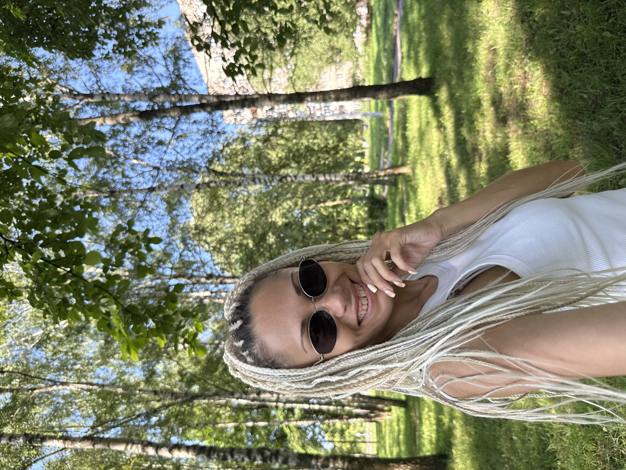 A woman with blonde braided hair and sunglasses smiles while resting her chin on her hand. She is wearing a white tank top. The woman is outdoors in a park with trees and green grass. In the background, there is a building and more trees. The scene appears to be located in Rusanovo, Russia, judging by the architecture and vegetation.