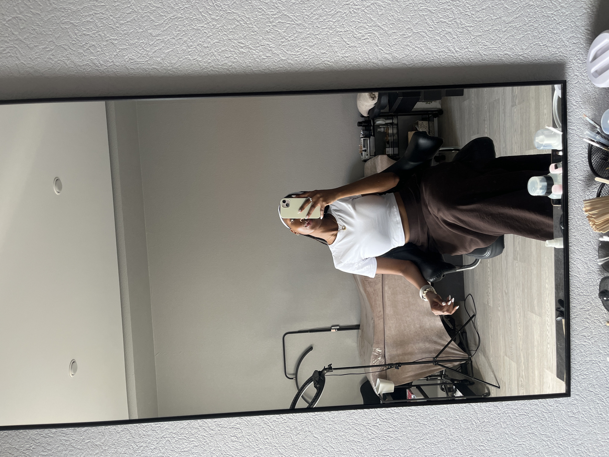 The image shows a young woman with dark skin and braids taking a selfie in a mirror. She is wearing a white crop top and a long brown skirt. She is seated in a black chair in what appears to be a studio or salon. In the background, reflected in the mirror, is a massage table covered with a tan sheet, a ring light on a tripod, and shelves with various products and supplies. The room has light grey walls and light-colored wood-look flooring. The overall setting suggests a professional beauty or wellness establishment. The location in Kazan, Russia is not specified by any visual cues in the image.