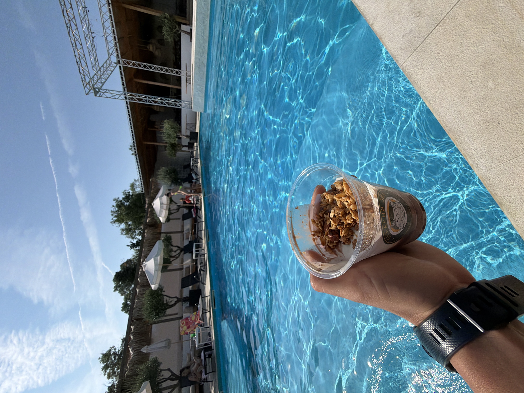 A person's hand, wearing a dark watch, holds a clear plastic cup of granola parfait near the edge of a vibrant blue swimming pool. The pool is situated outdoors, with lounge chairs, umbrellas, and lush green trees lining its perimeter. In the background, a covered structure with a metal framework is visible, suggesting an outdoor venue or entertainment space. The sky is a clear blue with wispy clouds. The image appears to be taken in Burgas, Bulgaria, specifically at a resort or beach club.