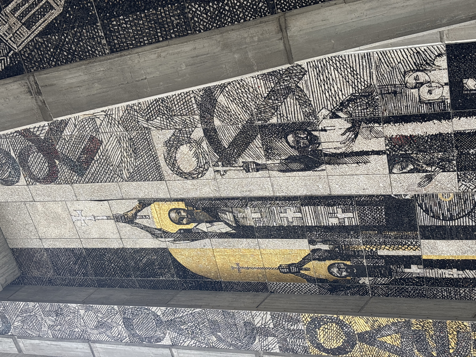 The media file showcases a large-scale mosaic artwork adorning a sloped ceiling or wall, framed by concrete architectural elements. The mosaic is rendered primarily in black and white tesserae, with significant use of gold and sparse red accents.
The artwork features stylized, iconographic figures. In the central horizontal band, a prominent figure with a gold halo and robes is depicted on the left, holding a staff or scepter topped with a cross-like finial. This figure is set against a background of white tesserae with intricate black hatching and cross-hatching patterns. Below this, a large, sweeping arc of solid gold tesserae forms a significant part of the background, beneath which another figure, partially visible and similarly styled, also holds a staff. Other sections of the mosaic display abstract geometric patterns, additional figurative elements, and discernible eyes or facial features integrated into complex designs.
The artistic style combines formal figure depiction with extensive use of detailed linear and hatched patterns to create texture and shading. The strategic use of gold highlights halos and significant background areas. The scene is static, presenting a formal composition of figures. No decipherable text is visible within the mosaic patterns. The concrete beams suggest a robust, possibly public or religious, architectural setting.