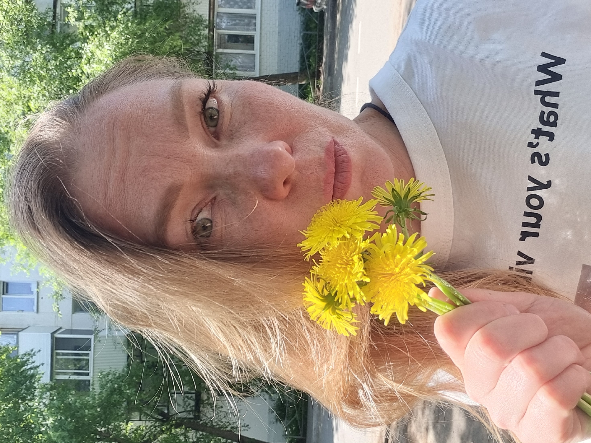 A woman is holding a bouquet of dandelions, seemingly in a city environment. The woman has fair skin, light blonde hair, and green eyes. She is wearing a white t-shirt with text printed on it. The bouquet consists of bright yellow dandelions held in her right hand. In the background, there's a multi-story residential building with windows. Trees are visible. The scene is likely in a city park or a residential area. Although I can not confirm this is specifically the city of Chaikovsky, Russia.