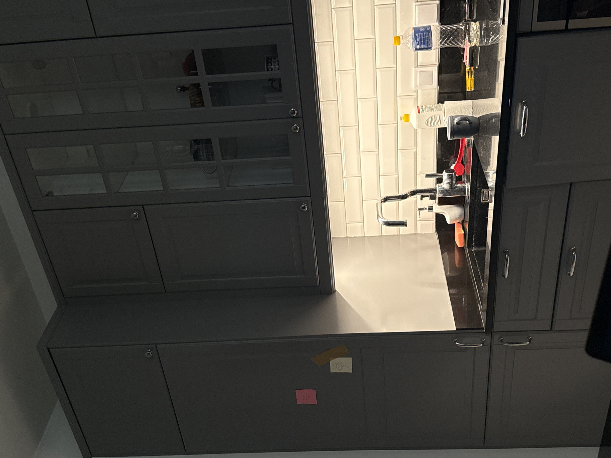 The image shows a modern kitchen counter with a sink, faucet, and various cleaning supplies. A bottle of water, dish soap, and a roll of paper towels are visible on the counter. There is also a dark-colored mug and a red sponge. Above the counter, there are white subway tiles and dark gray cabinets. The cabinets have glass doors in the upper section, revealing some items inside. Two sticky notes are attached to the cabinet door on the left. The lighting is dim, with under-cabinet lights illuminating the counter space. The overall scene suggests a clean and organized kitchen environment.