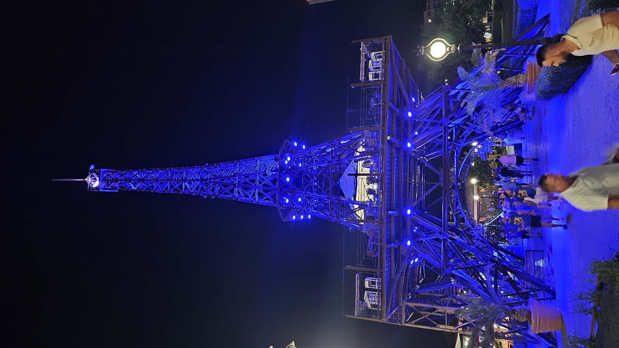 The image shows a nighttime view of a replica of the Eiffel Tower, illuminated with blue lights, located in the city of Varna, Bulgaria. The tower is a prominent structure in the center of the frame, with its intricate metal framework clearly visible. Several levels of the tower are lit up, and there appear to be tables and chairs set up on one of the upper platforms, suggesting a seating area or restaurant.
At the base of the tower, a pathway is visible, with several people walking. The path is also bathed in blue light. To the left, a large potted palm tree stands near the base. To the right, a decorative lamppost with a glowing orb illuminates the scene. In the background, trees and buildings are faintly visible against the dark night sky. The overall scene suggests an entertainment or recreational area in Varna, likely a popular spot for visitors and locals alike.