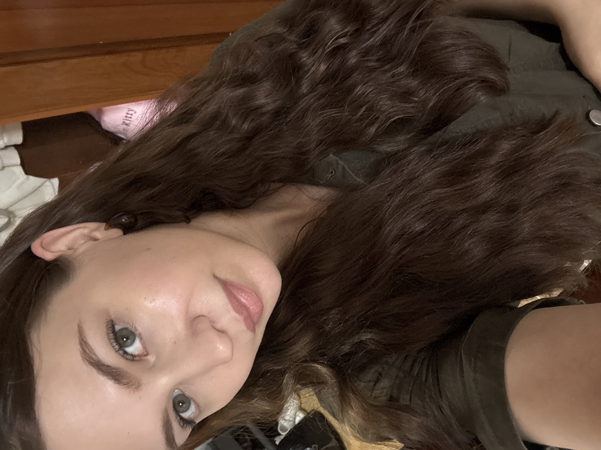 The image shows a close-up, selfie-style shot of a young Caucasian woman with long, wavy brown hair. She is wearing a dark green collared shirt, possibly a button-down. Her skin appears smooth and lightly made up, with a subtle sheen on her cheek. She has green eyes and her eyebrows are well-defined. Her lips are glossed with a pinkish-nude shade. She is wearing a dark, possibly tortoiseshell, earring in her visible ear. In the background, to the right, a portion of a wooden surface, likely a door frame or furniture, is visible. Behind her left ear, a pink garment with "Kitty" printed in a darker font is partially visible. The lighting suggests an indoor setting, possibly a room in a residential area. There is no visual information in the image that can be used to determine its location within the city of Chaikovsky, Russia.
