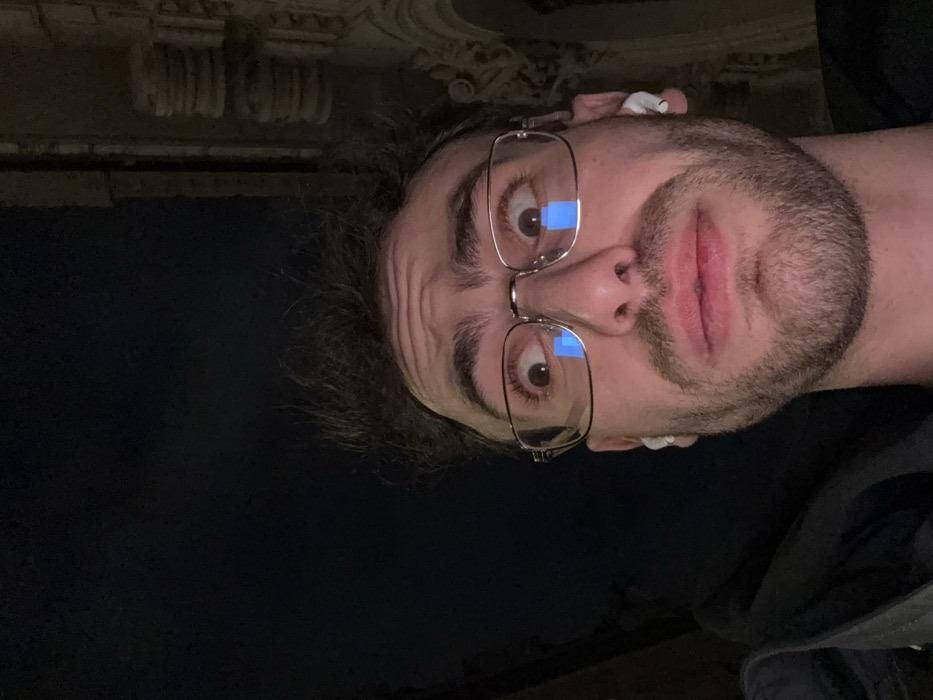 A man with dark hair, a beard, and glasses takes a selfie. He is wearing wireless earbuds. His face is close to the camera, and he looks surprised or shocked. The background is dark, but there is an ornate, upside-down design visible at the top of the frame, suggesting he is indoors. The image is tilted, making the man appear sideways.